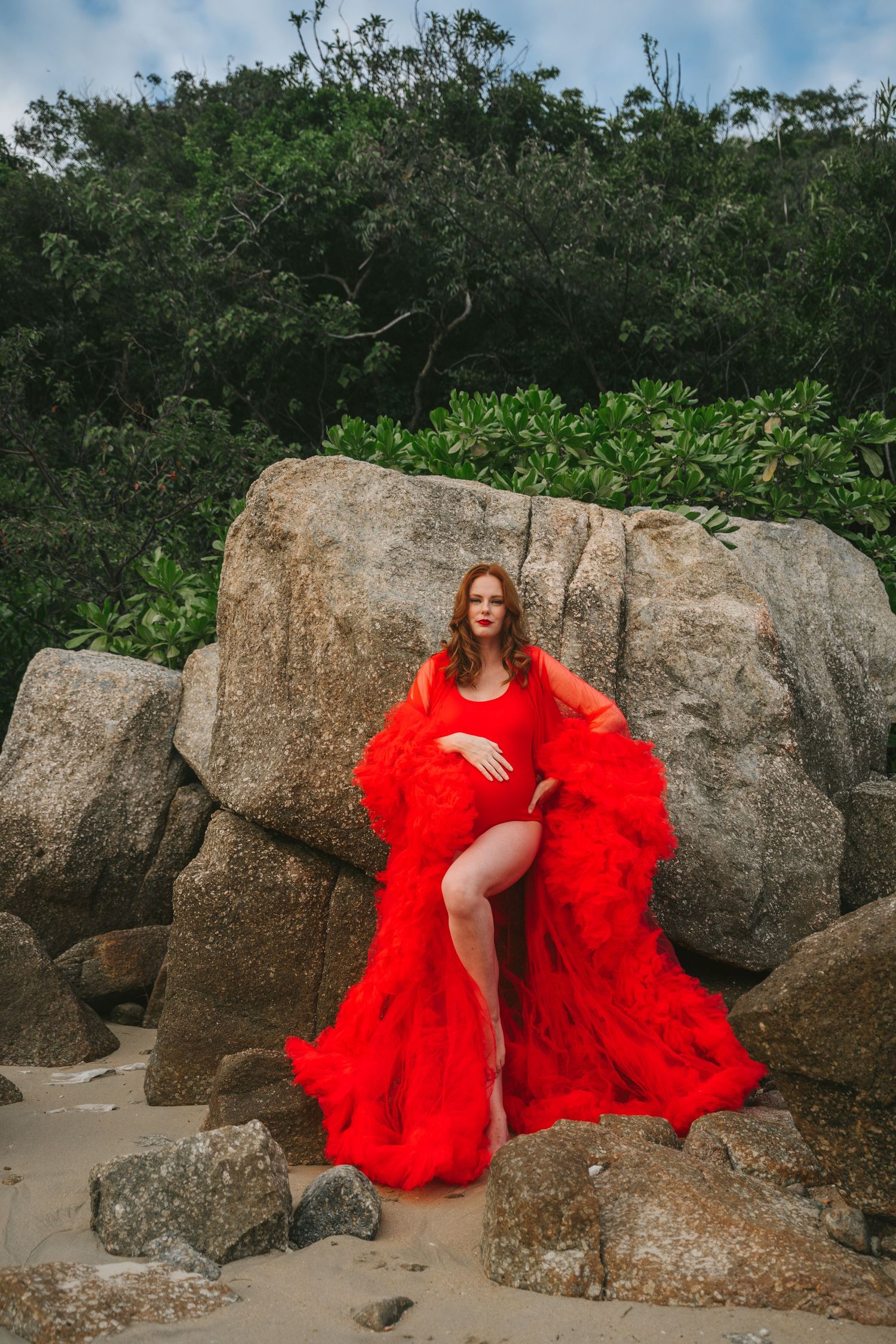 Fashion portrait on rocky beach with flowing red dress against tropical foliage.
