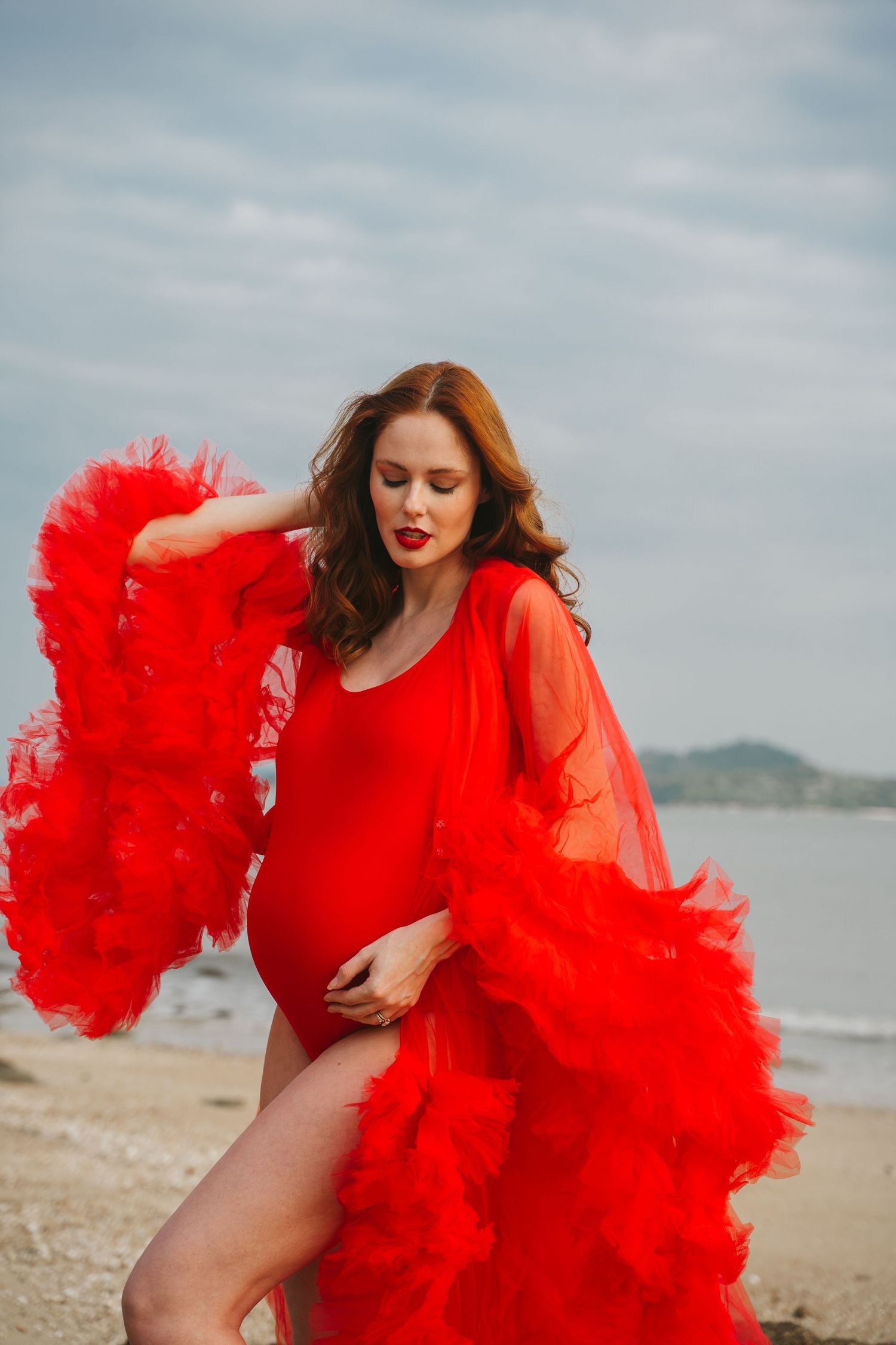 Dramatic maternity photo with flowing red dress against coastal backdrop.
