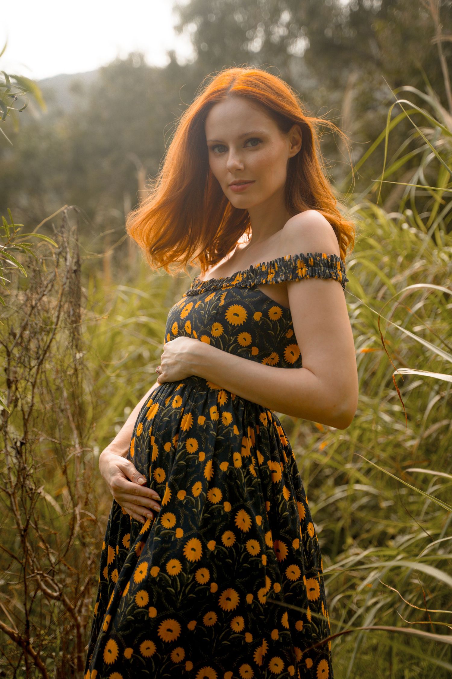 Expecting mother in floral dress poses in sunlit natural setting.