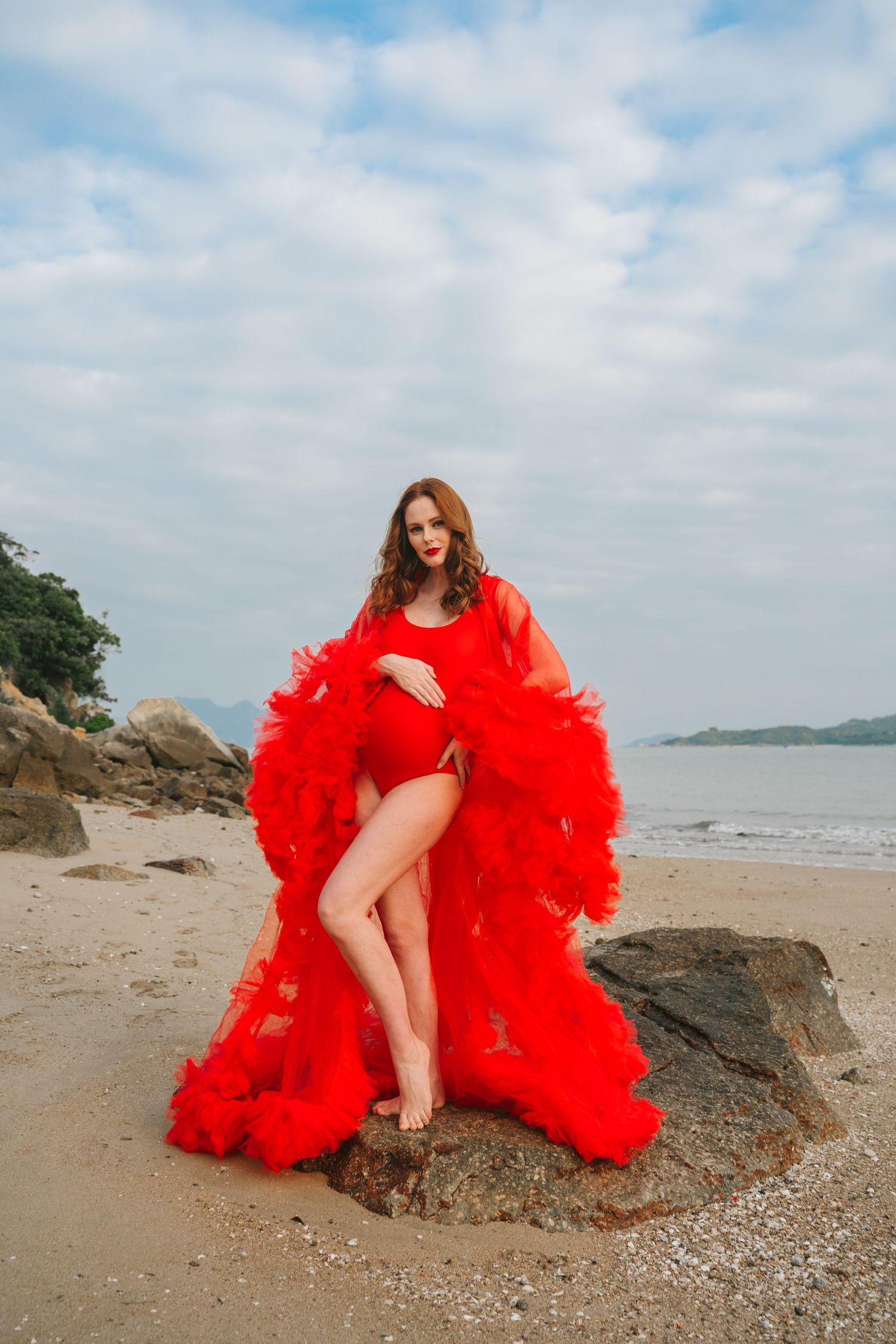 Model in vibrant red swimsuit and dramatic tulle robe poses on coastal rocks.