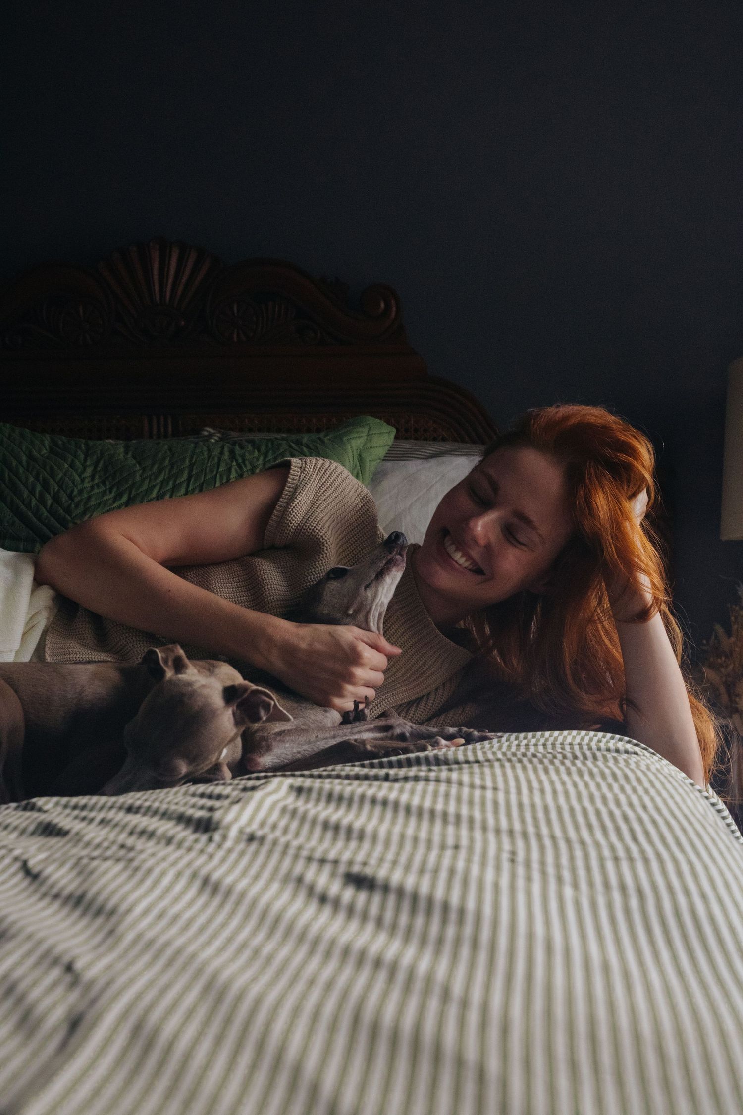 Woman bonding with Italian Greyhound while relaxing on bed in dimly lit room.