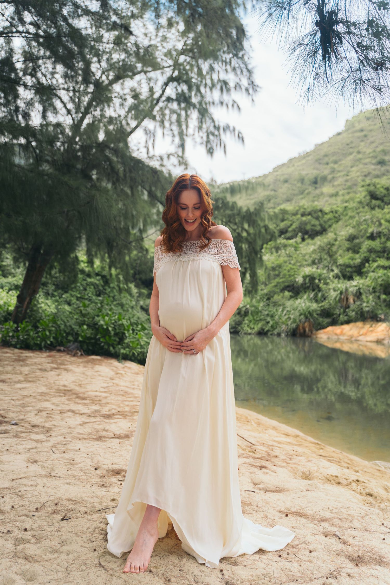 Beachside maternity photo featuring flowing white dress and mountain scenery.