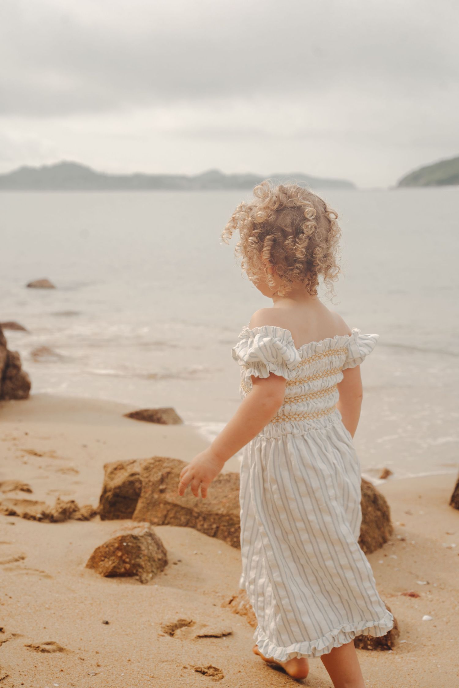 A small child in a striped dress explores a rocky beach on an overcast day.