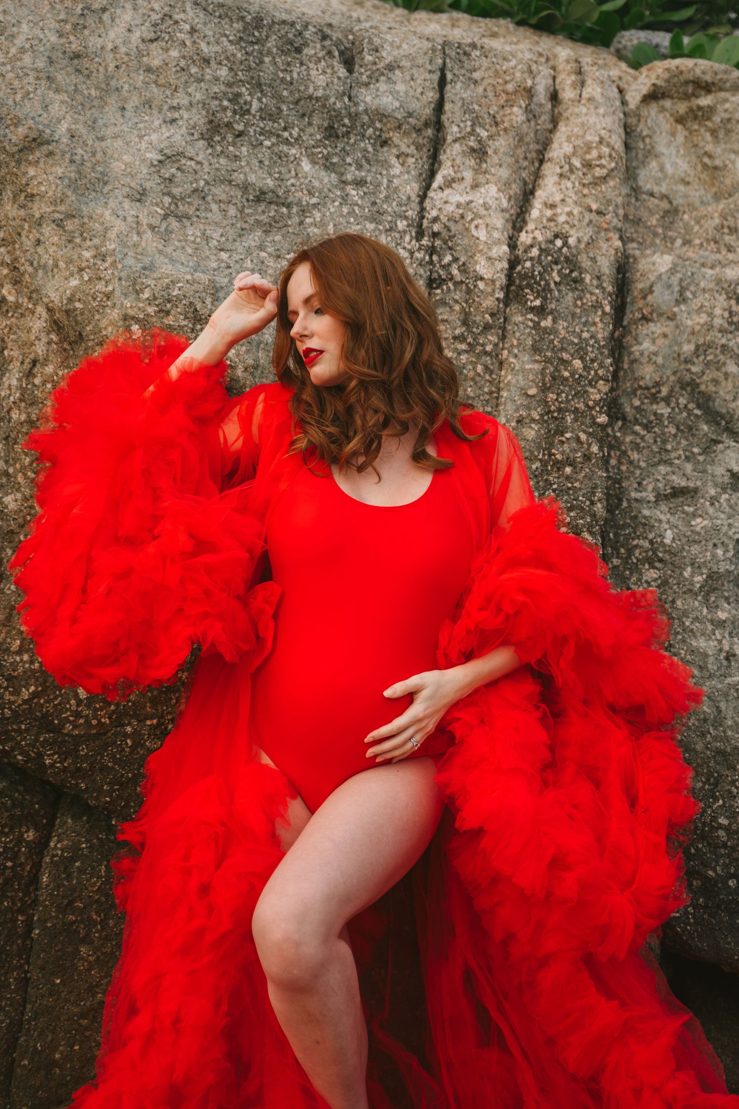 Dramatic red ruffled dress contrasts against rocky backdrop in artistic maternity photo.