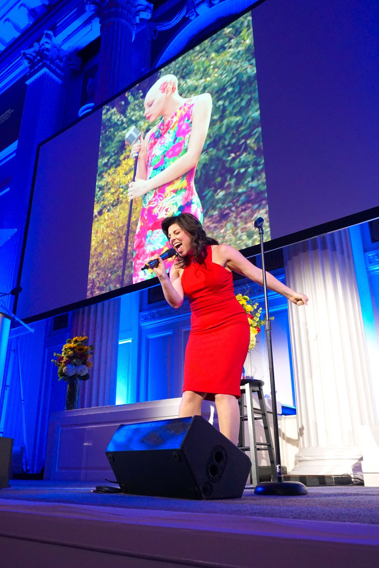 A performer in a red dress sings on stage at an event with dramatic blue lighting and a large screen backdrop.