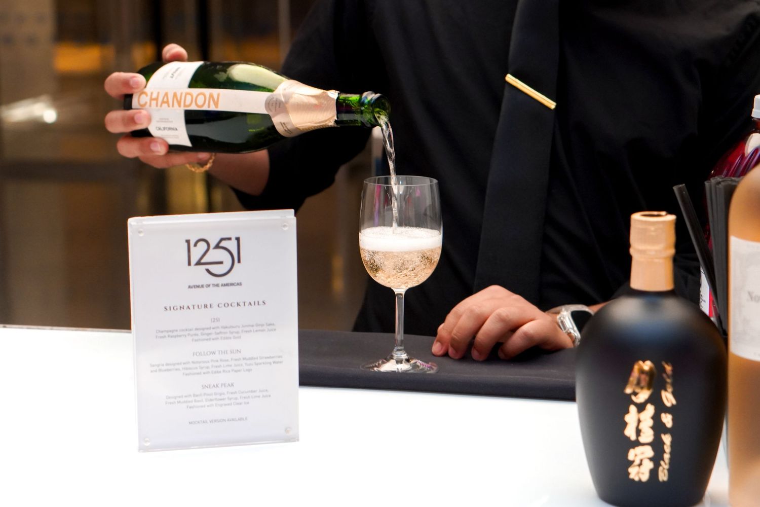 A bartender pours a drink at an event with a '1251' menu card displayed on the table.