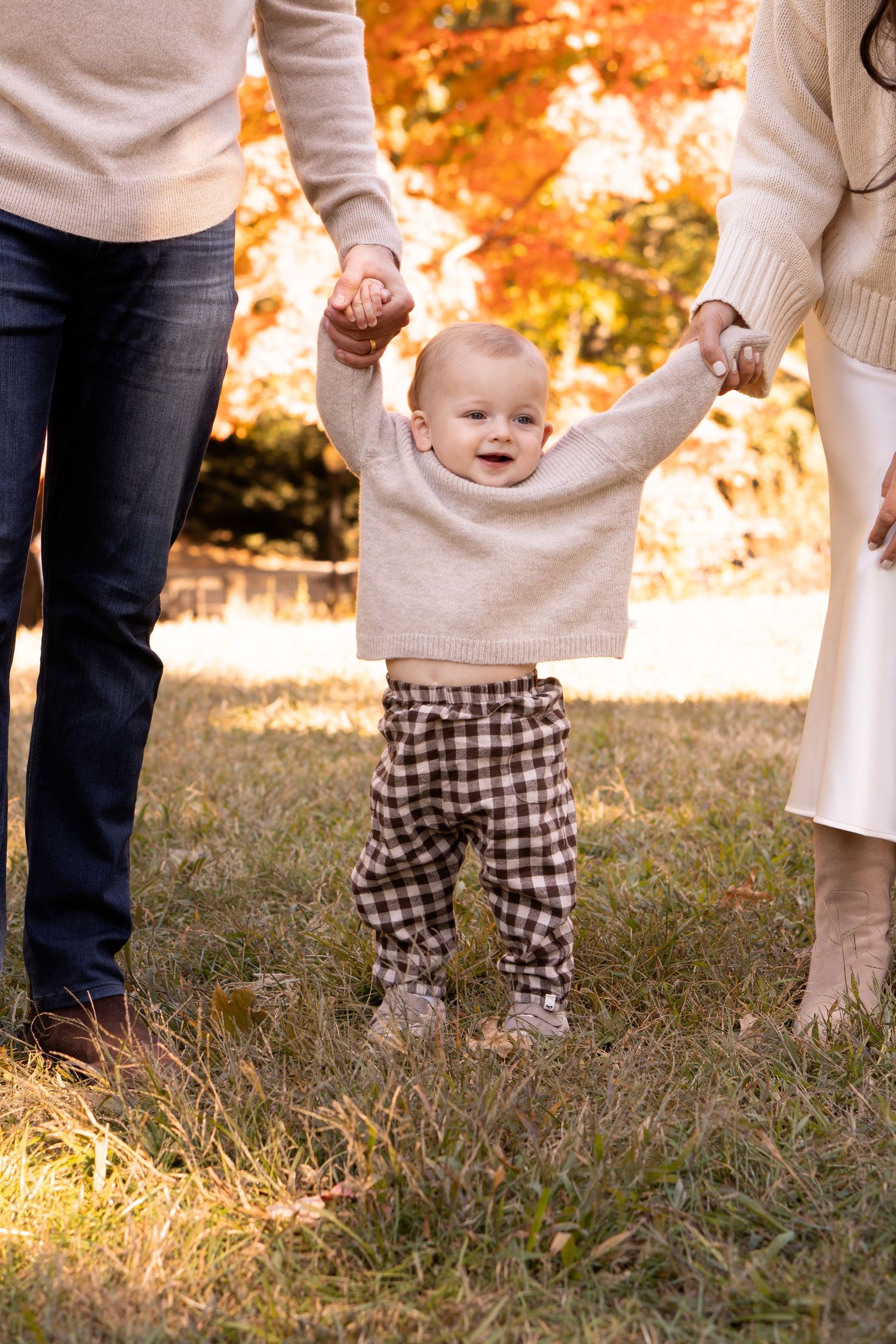 Smiling baby in beige sweater walking with parents amid fall foliage.