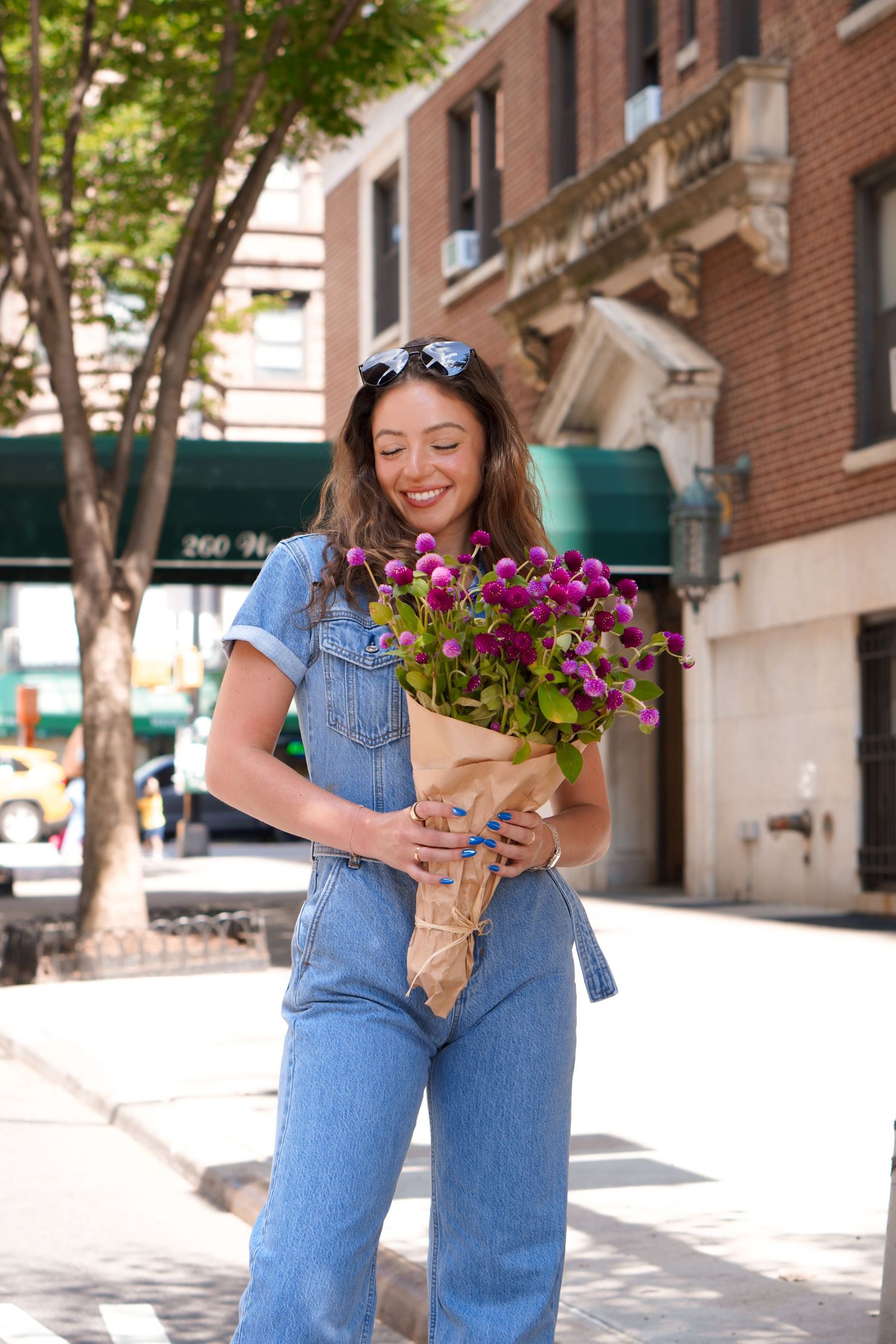 Person carrying fresh purple flowers in denim overalls on an urban sidewalk.