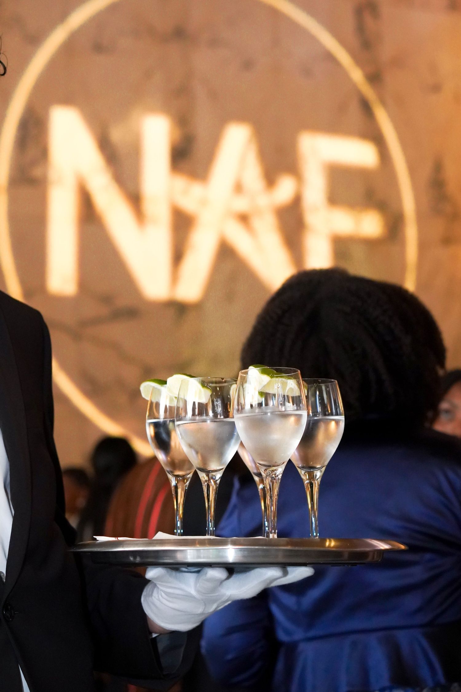 Server carrying champagne glasses on a tray with illuminated NAF logo in background.