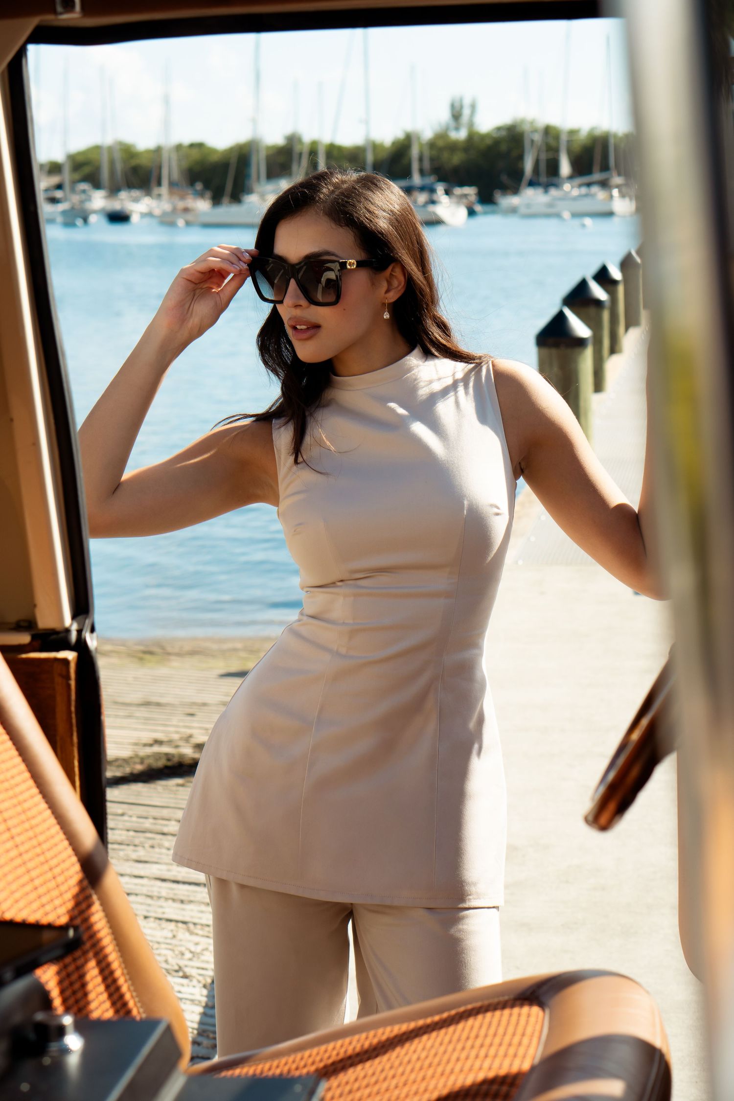 Woman in beige sleeveless outfit and sunglasses posing at a sunny marina with sailboats in the background.