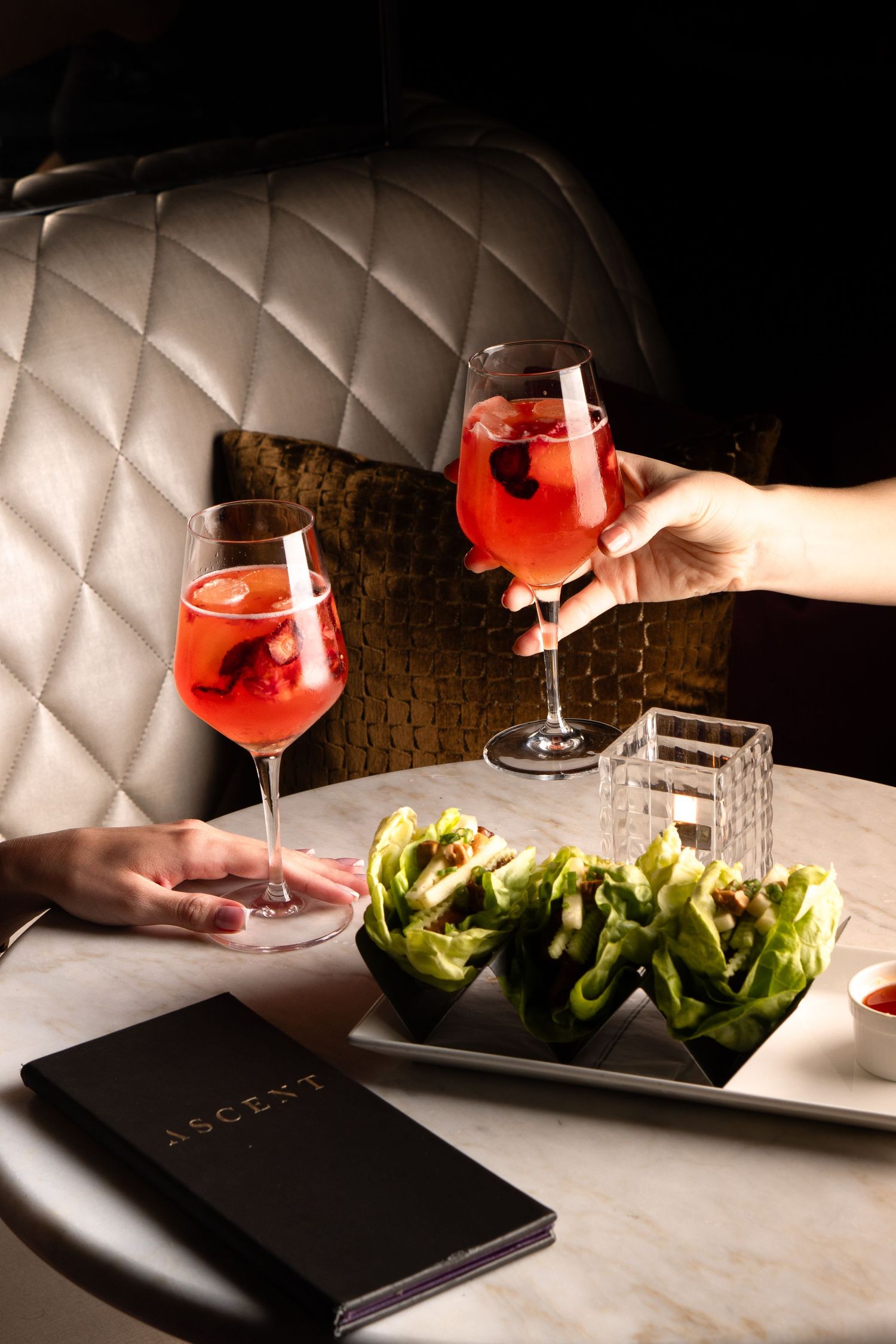 Two spritz cocktails and fresh lettuce cups on a quilted banquette in a dimly lit restaurant setting.