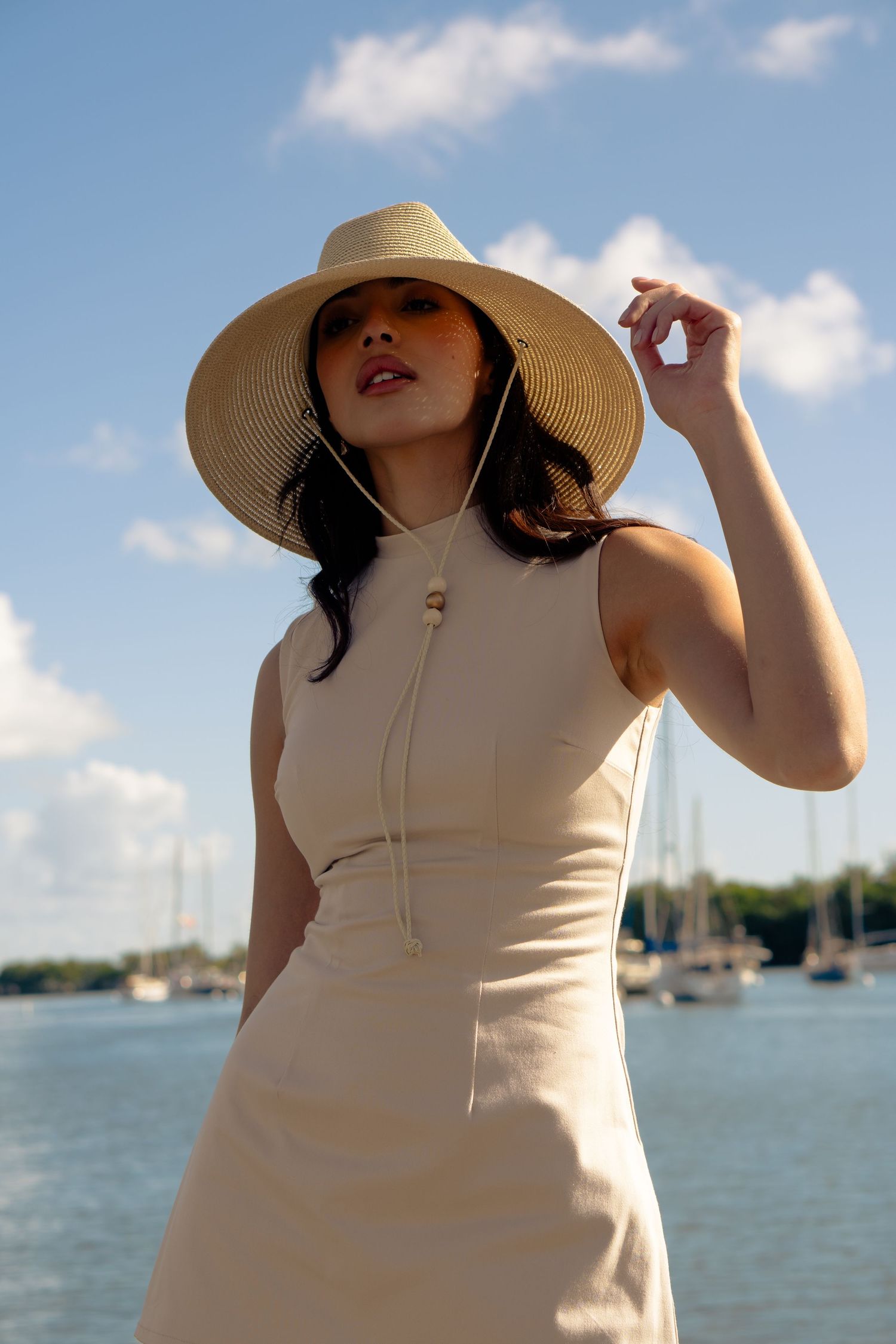 Woman in beige sleeveless dress and wide-brim straw hat poses near a marina with sailboats and blue sky.