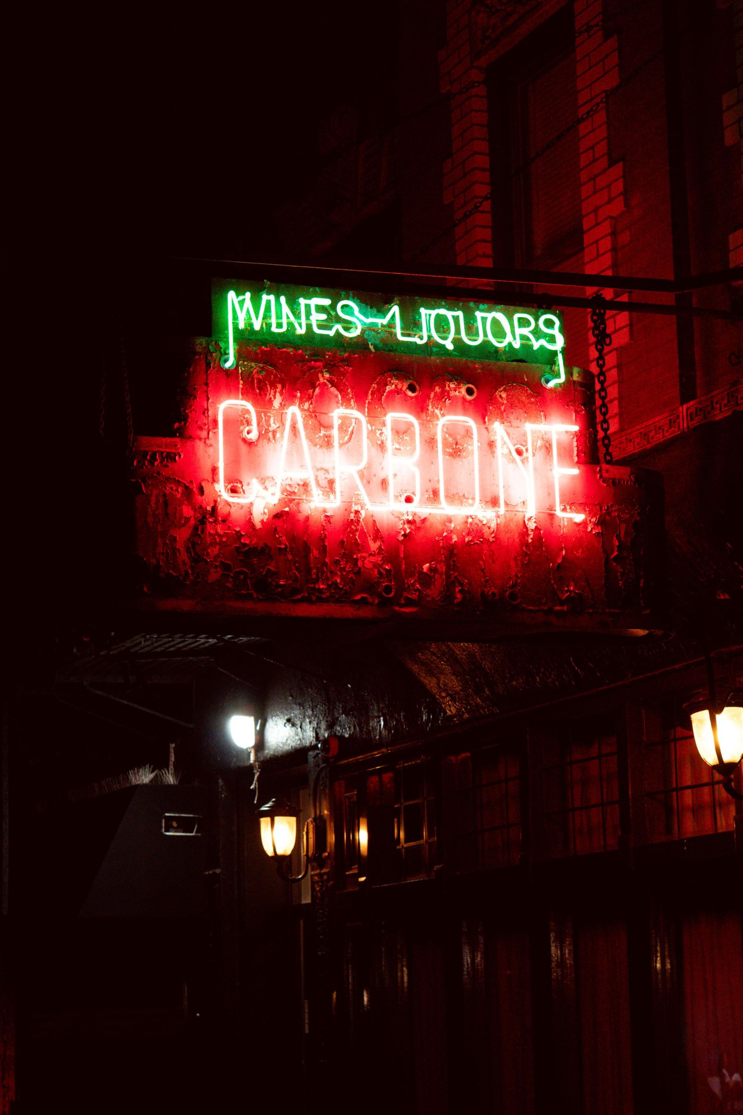 Neon signs for Carbone restaurant and Wines & Liquors glow red and green against a dark brick building at night.