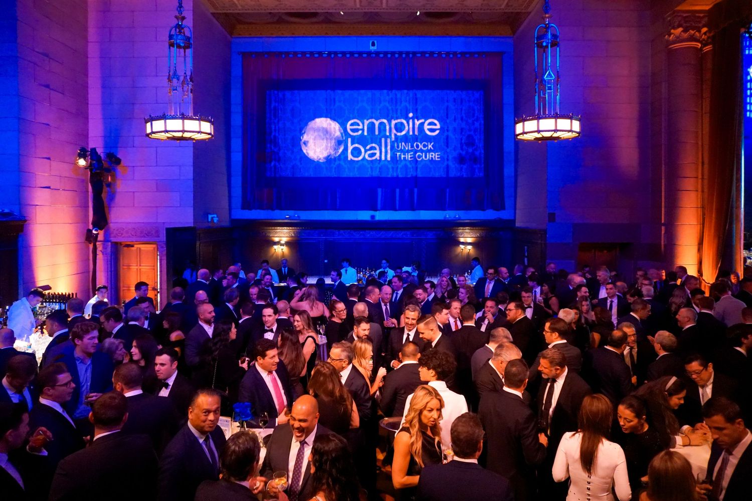Crowded Empire Ball gala event with guests in formal attire gathered under blue illuminated signage.