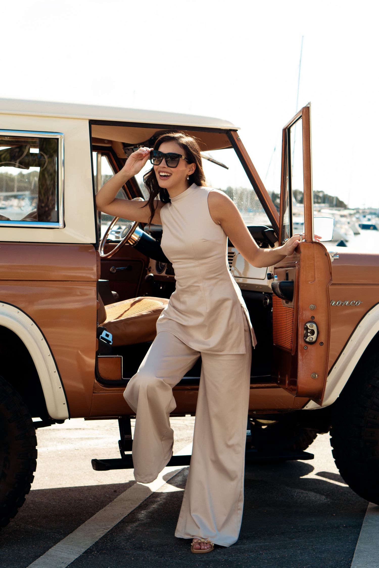Woman in beige wide-leg jumpsuit posing beside a vintage brown Ford Bronco, wearing sunglasses outdoors.