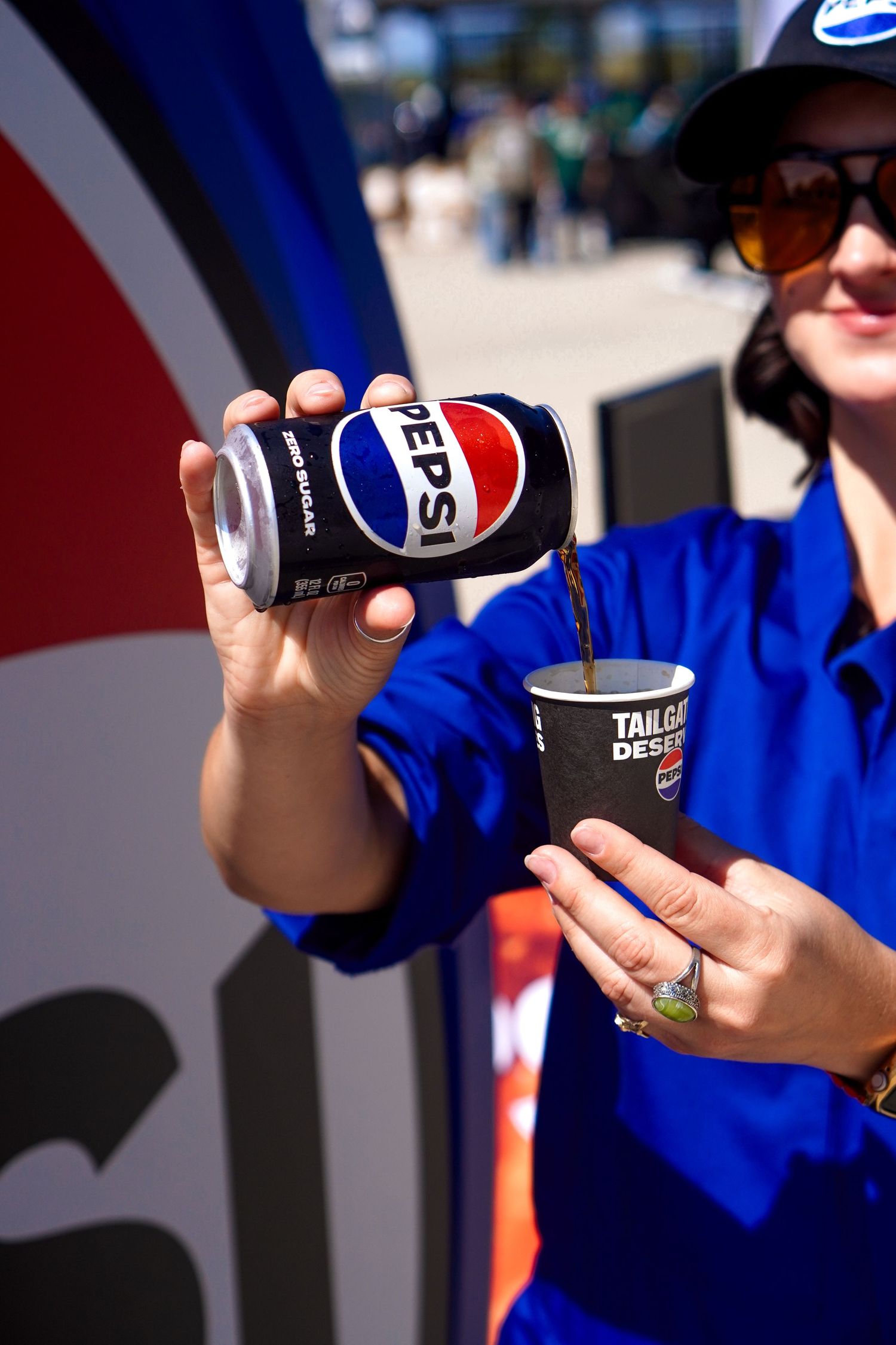 A Pepsi employee in blue uniform pours a can of Pepsi into a cup at a tailgate event outdoors.