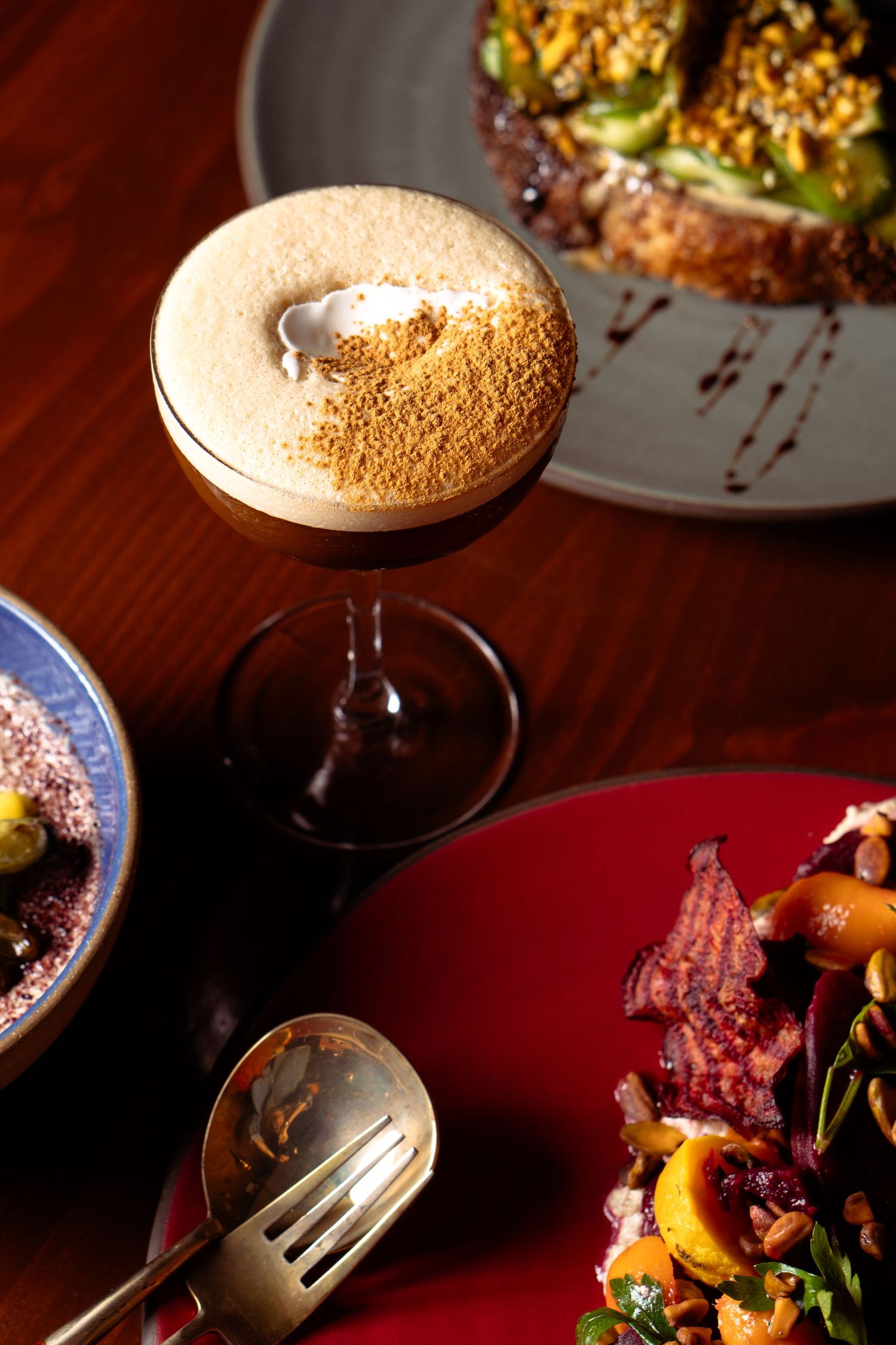 Espresso martini with foam topping surrounded by gourmet dishes on a wooden table.