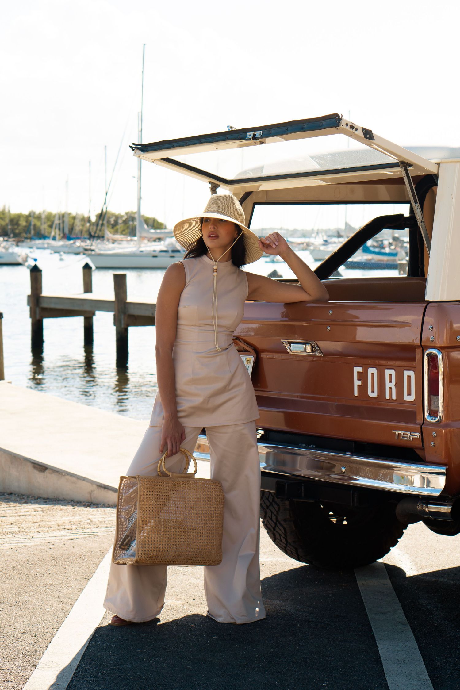 Woman in beige outfit and sun hat poses beside a vintage brown Ford Bronco at a marina dock.