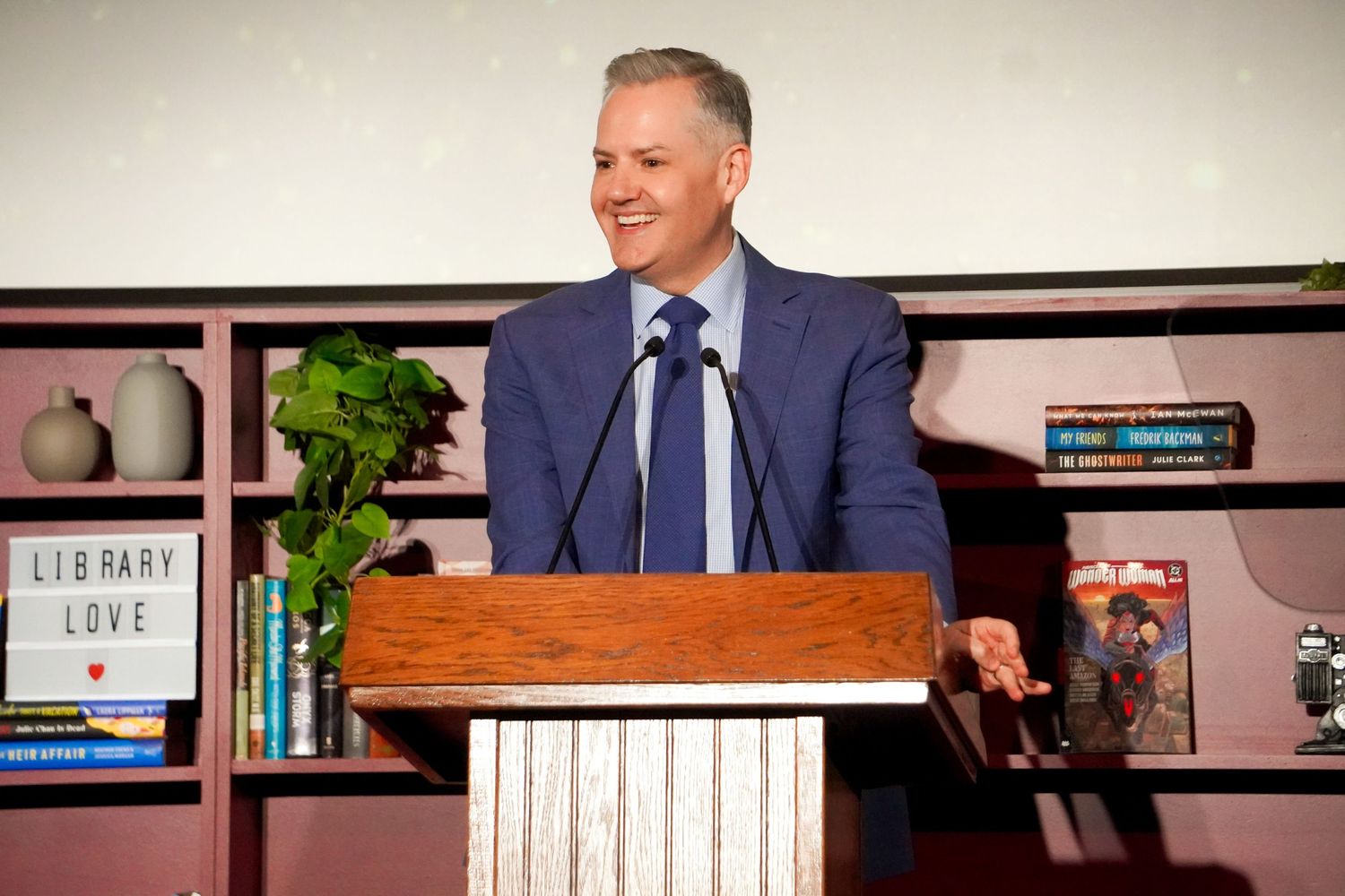 Man in blue suit speaking at wooden podium near bookshelf with 'Library Love' sign at formal event.