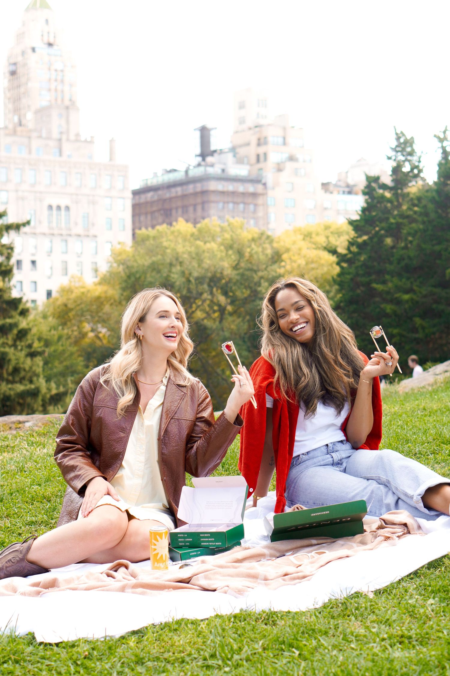 Friends laughing and enjoying drinks during outdoor picnic with city skyline behind them.