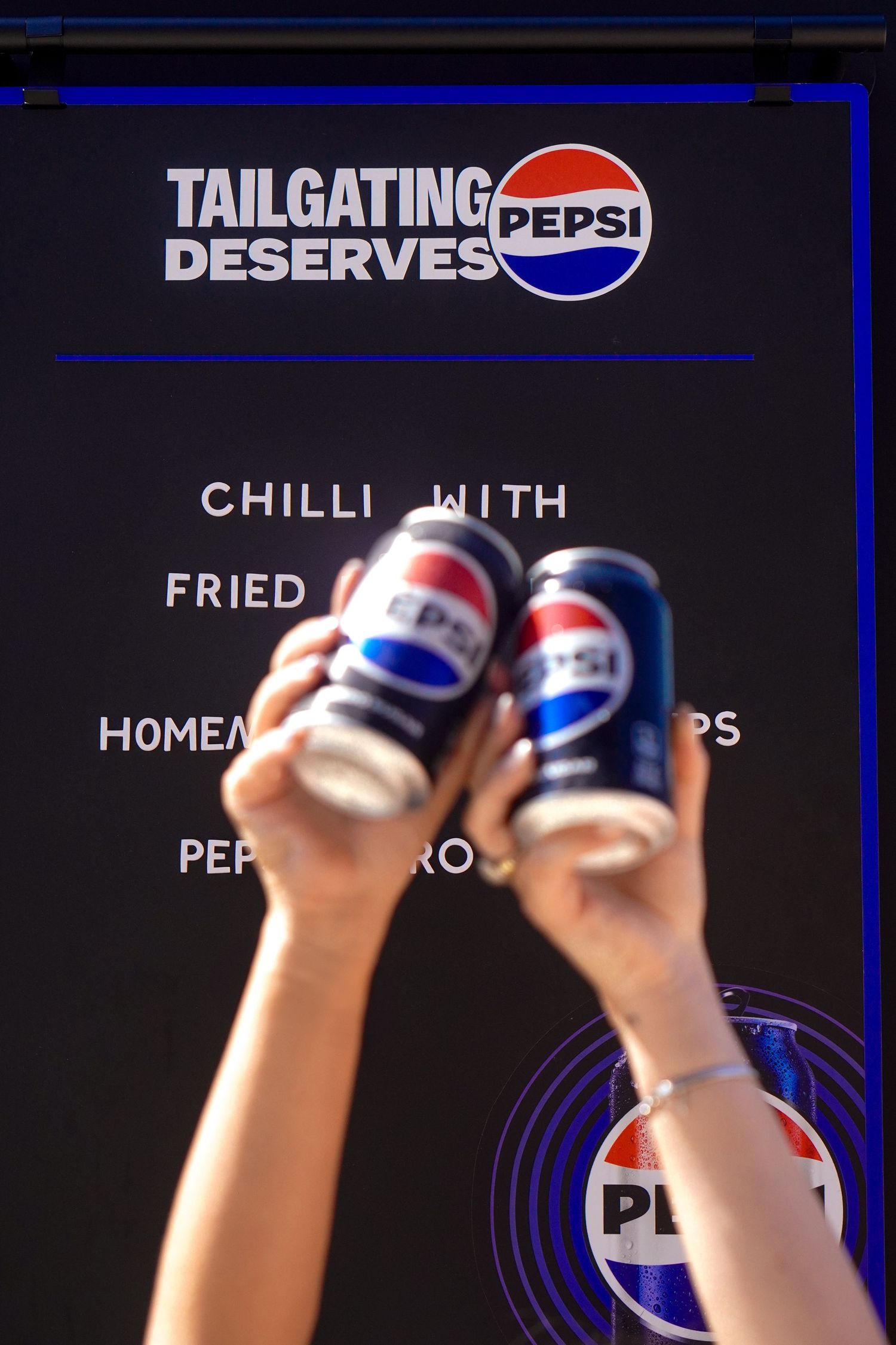 Two hands toasting Pepsi cans in front of a Tailgating Deserves Pepsi branded menu sign.