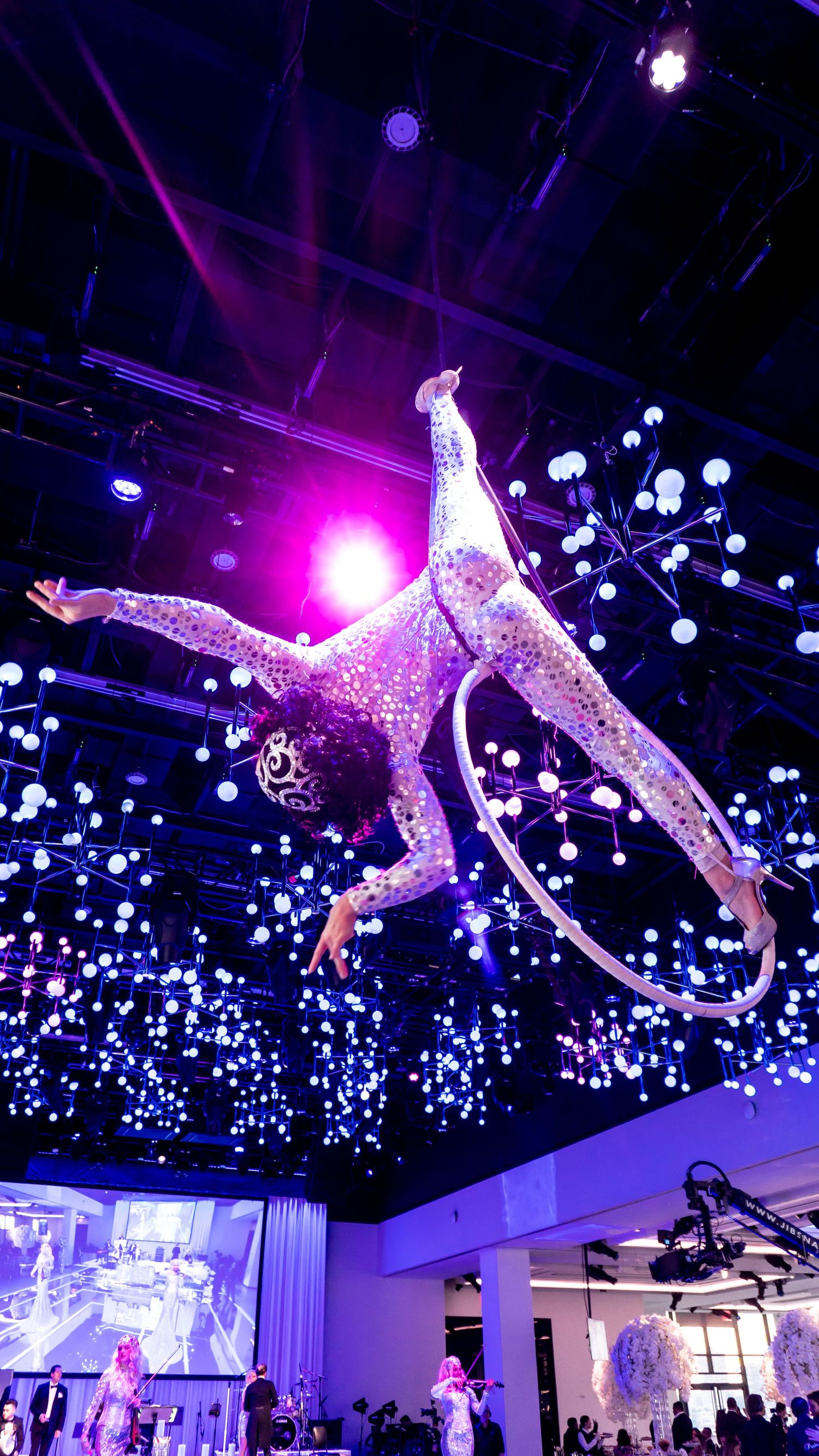 Aerial performer in glittering costume suspended amid purple lighting and illuminated spheres.