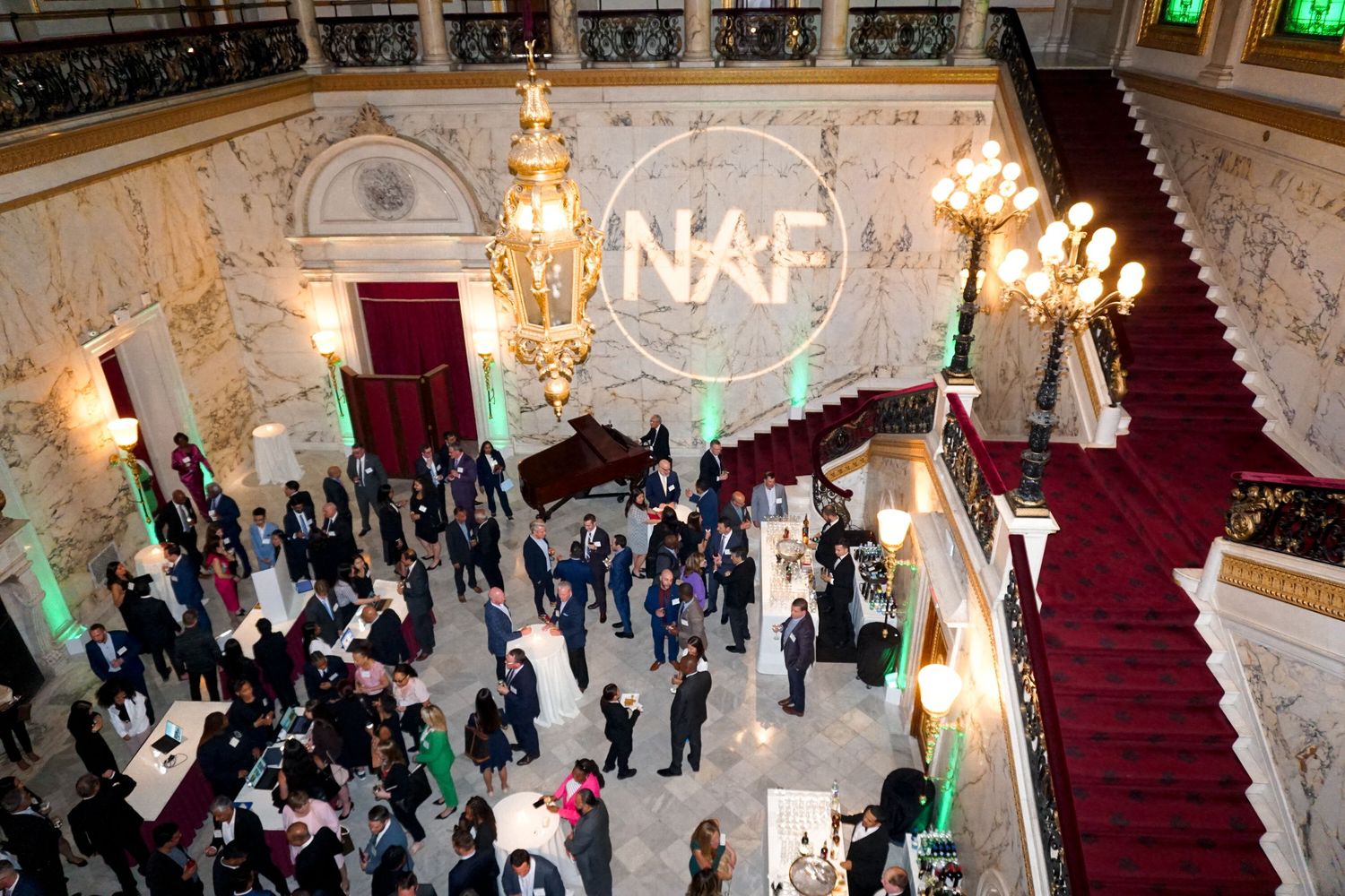 Aerial view of formal event with guests mingling under crystal chandeliers and NAF logo projection.