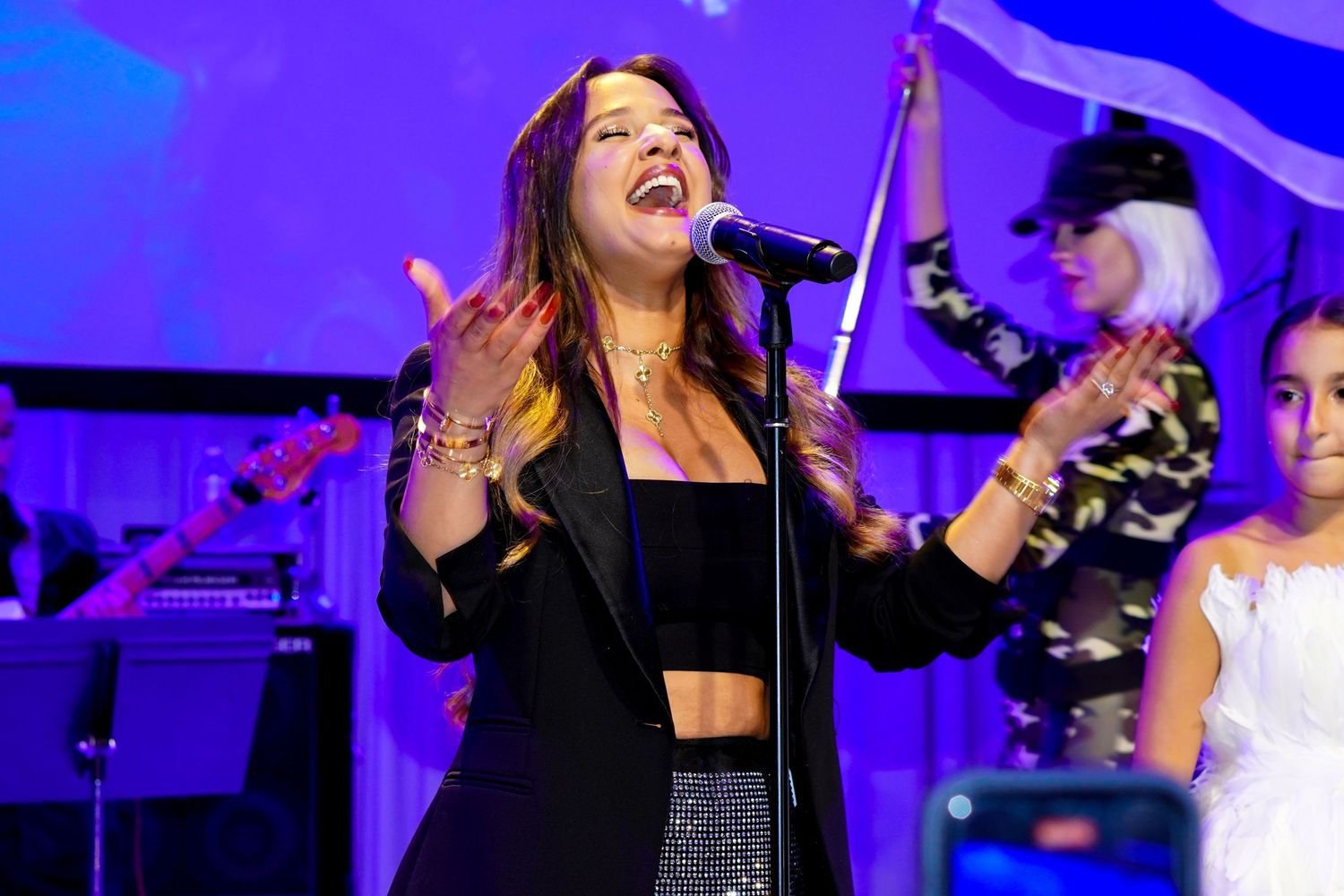 Female singer performing energetically at an event with blue lighting and musicians in background.
