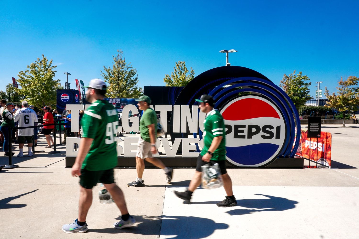 NFL fans walk past a large Pepsi Tailgating Deserves branded display outside a stadium on a sunny day.