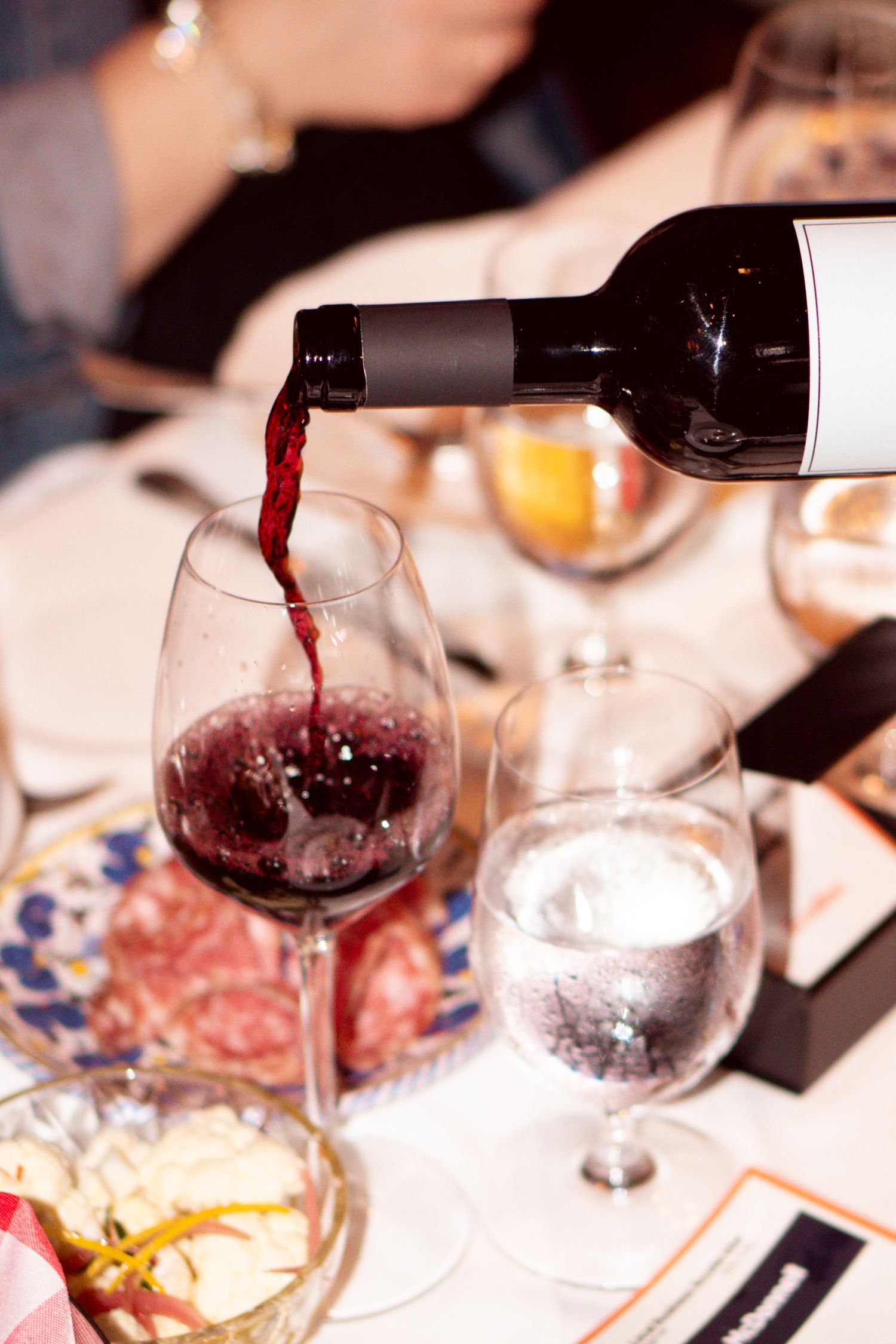Red wine being poured into a glass at a dinner table with charcuterie board and water glass.