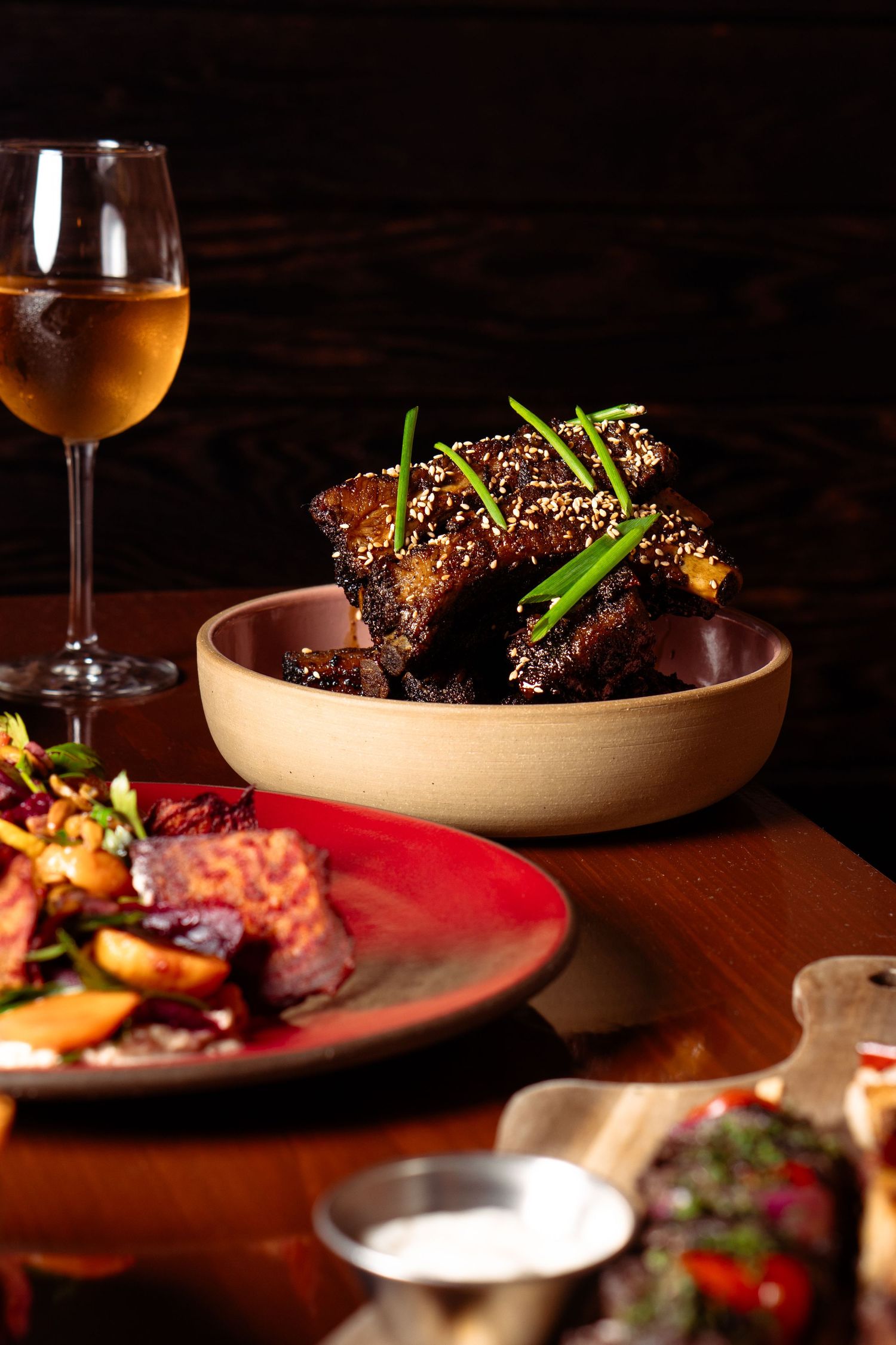 Glazed ribs topped with sesame seeds and green onions in a bowl, served with roasted vegetables and white wine.