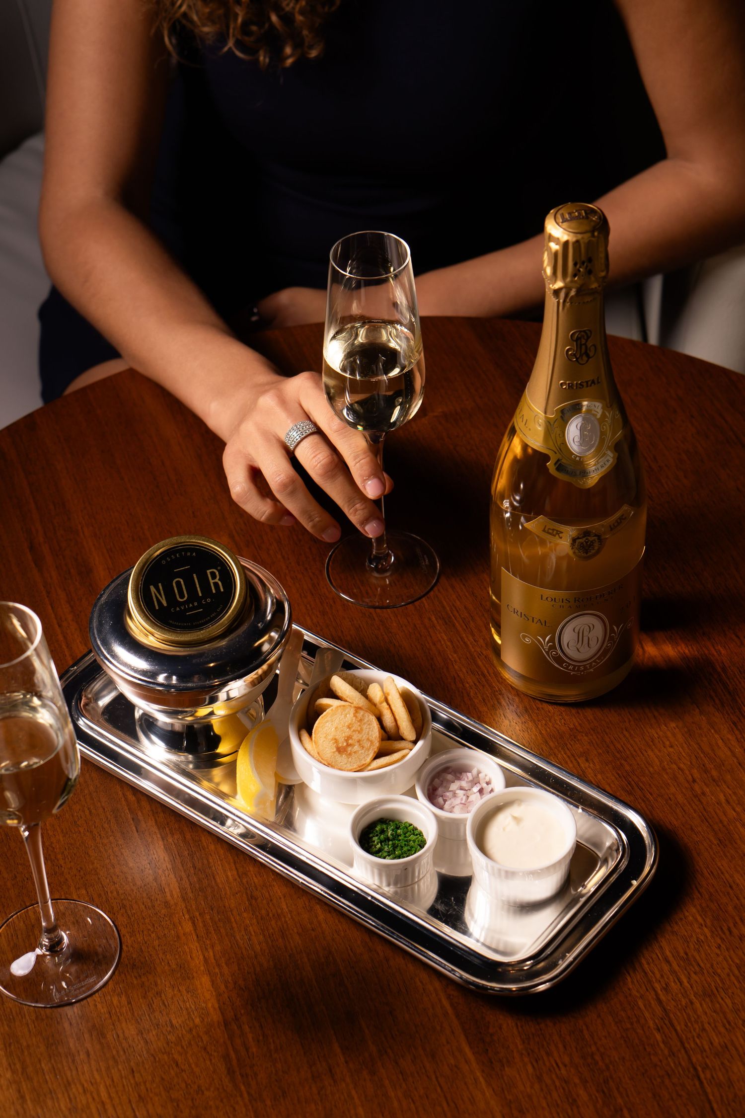 Woman holding champagne glass with Cristal bottle, caviar tin, and elegant appetizer tray on wooden table.