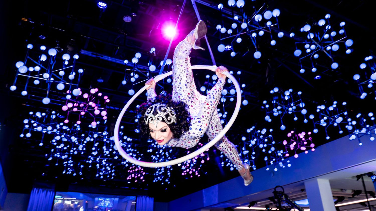 Aerial performer in glittering costume suspended beneath blue-lit ceiling with twinkling lights.