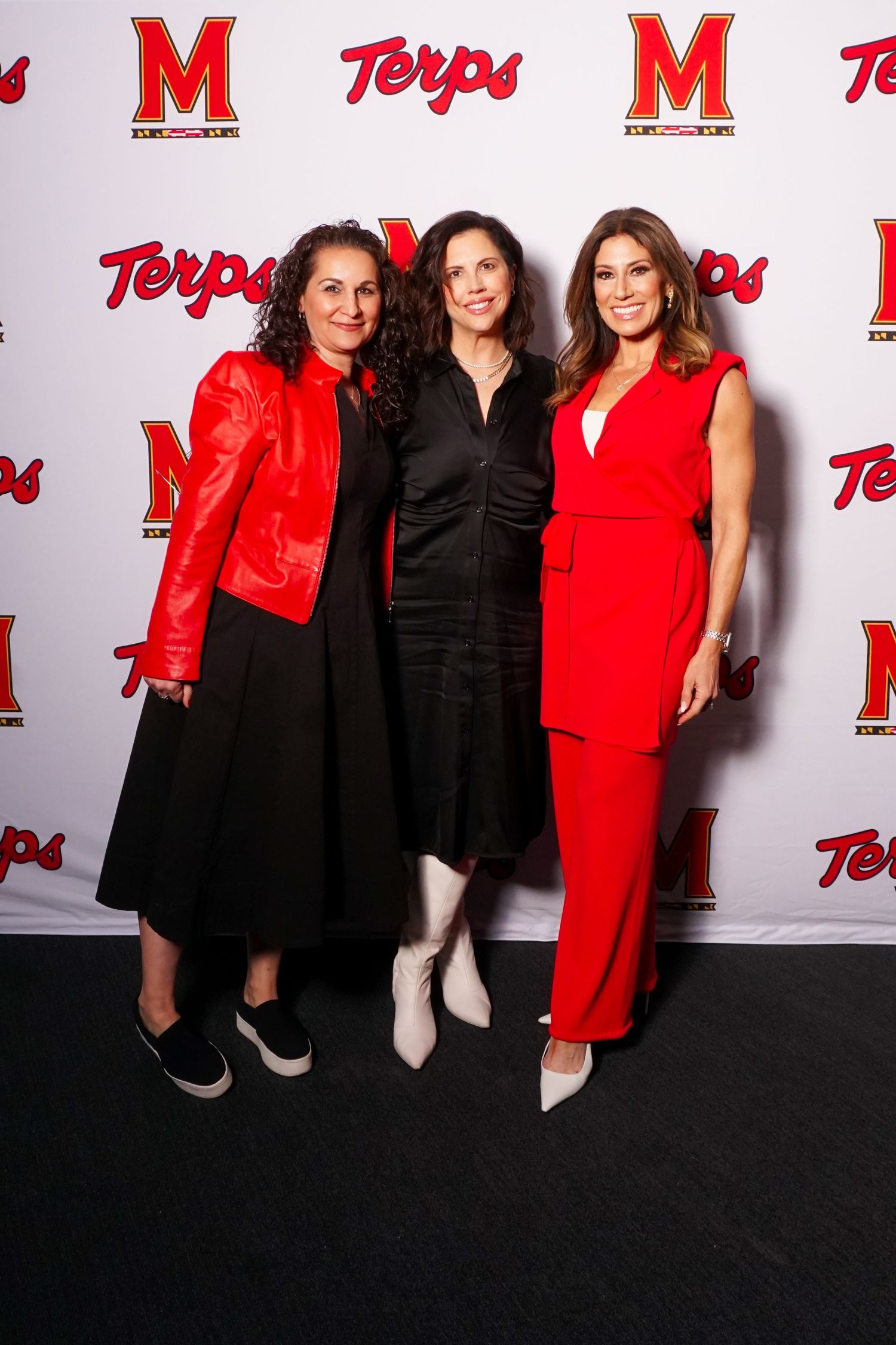 Three women pose together at a Maryland Terps event, two in red outfits and one in black, against a branded backdrop.