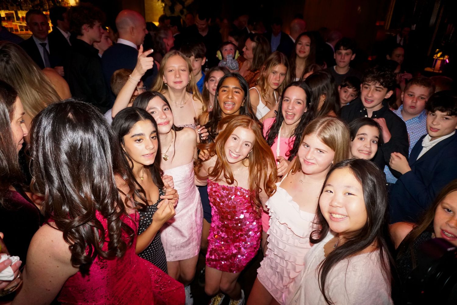 A large group of teens in formal attire smile and pose together at a party or school dance event.