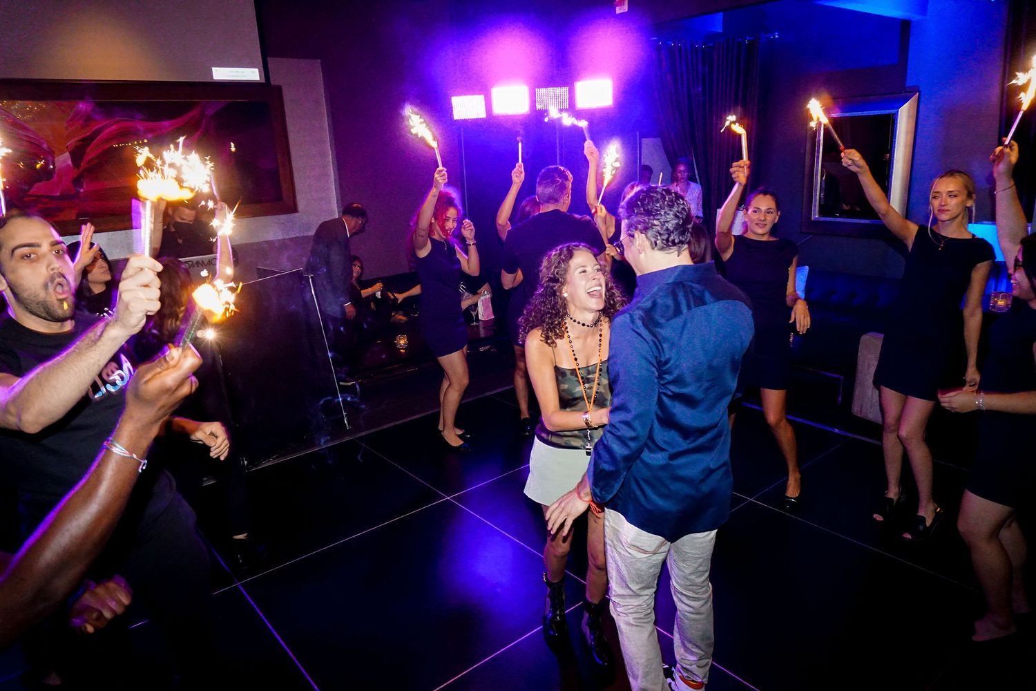 People dancing and celebrating with sparklers in a darkened venue with purple lighting.