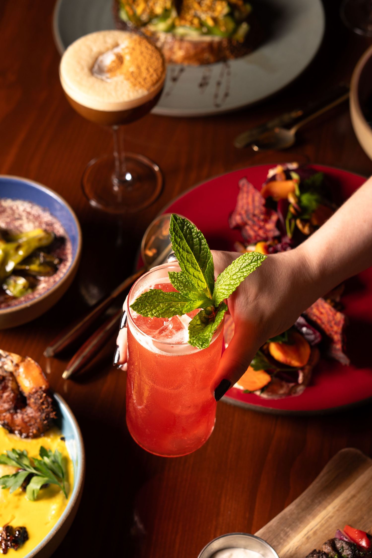 Hand holding a pink cocktail garnished with mint leaves, surrounded by colorful dishes on a wooden table.