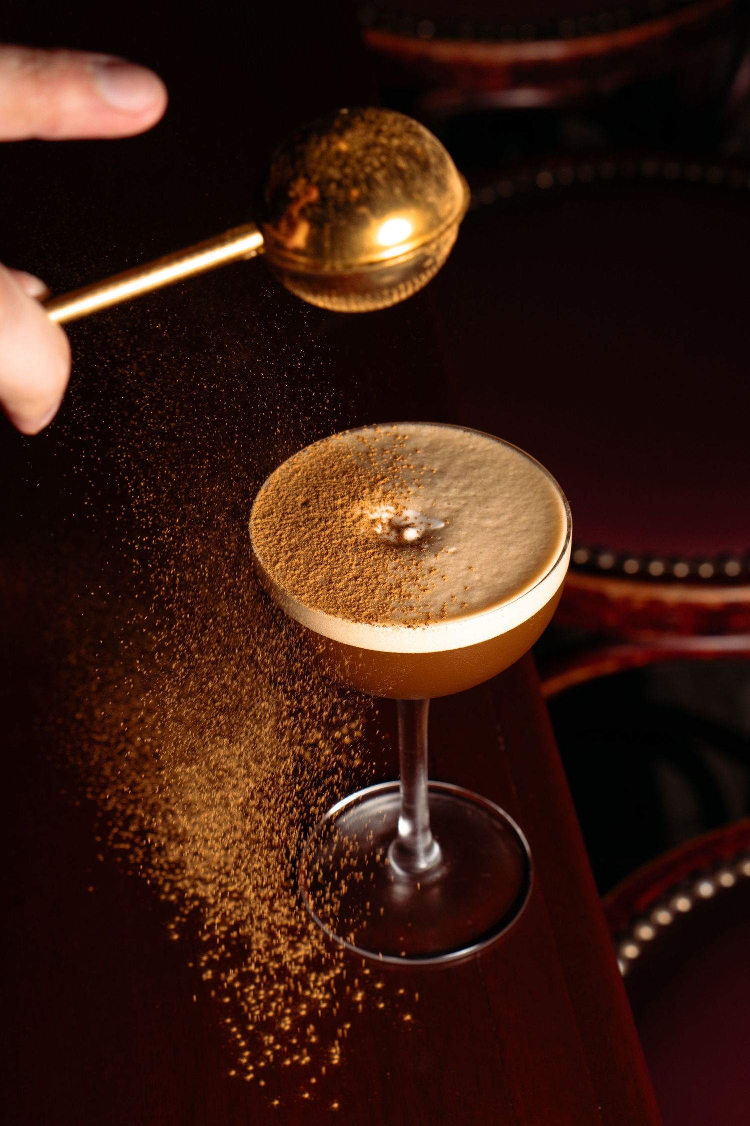 Bartender sprinkles cocoa powder over an espresso martini cocktail in a coupe glass on a dark wooden surface.
