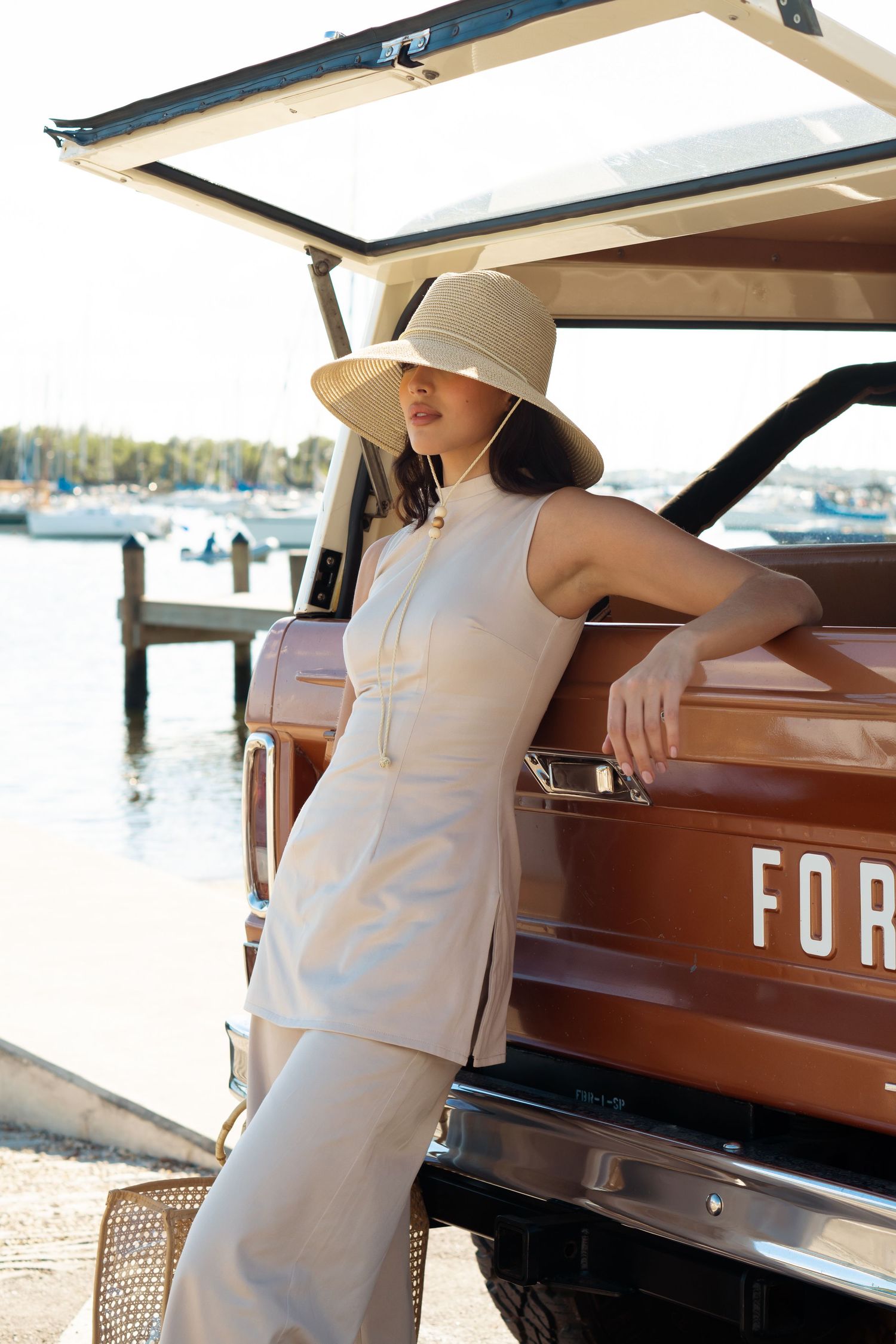 Woman in beige outfit and sun hat leaning against a vintage Ford SUV near a marina on a sunny day.
