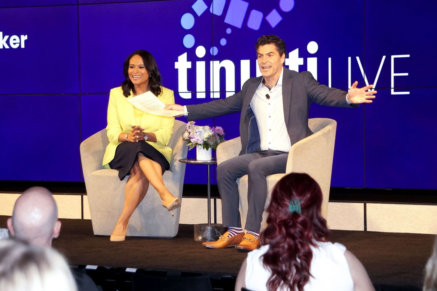 Conference speakers seated in armchairs on stage at Tinuiti Live event with blue backdrop.
