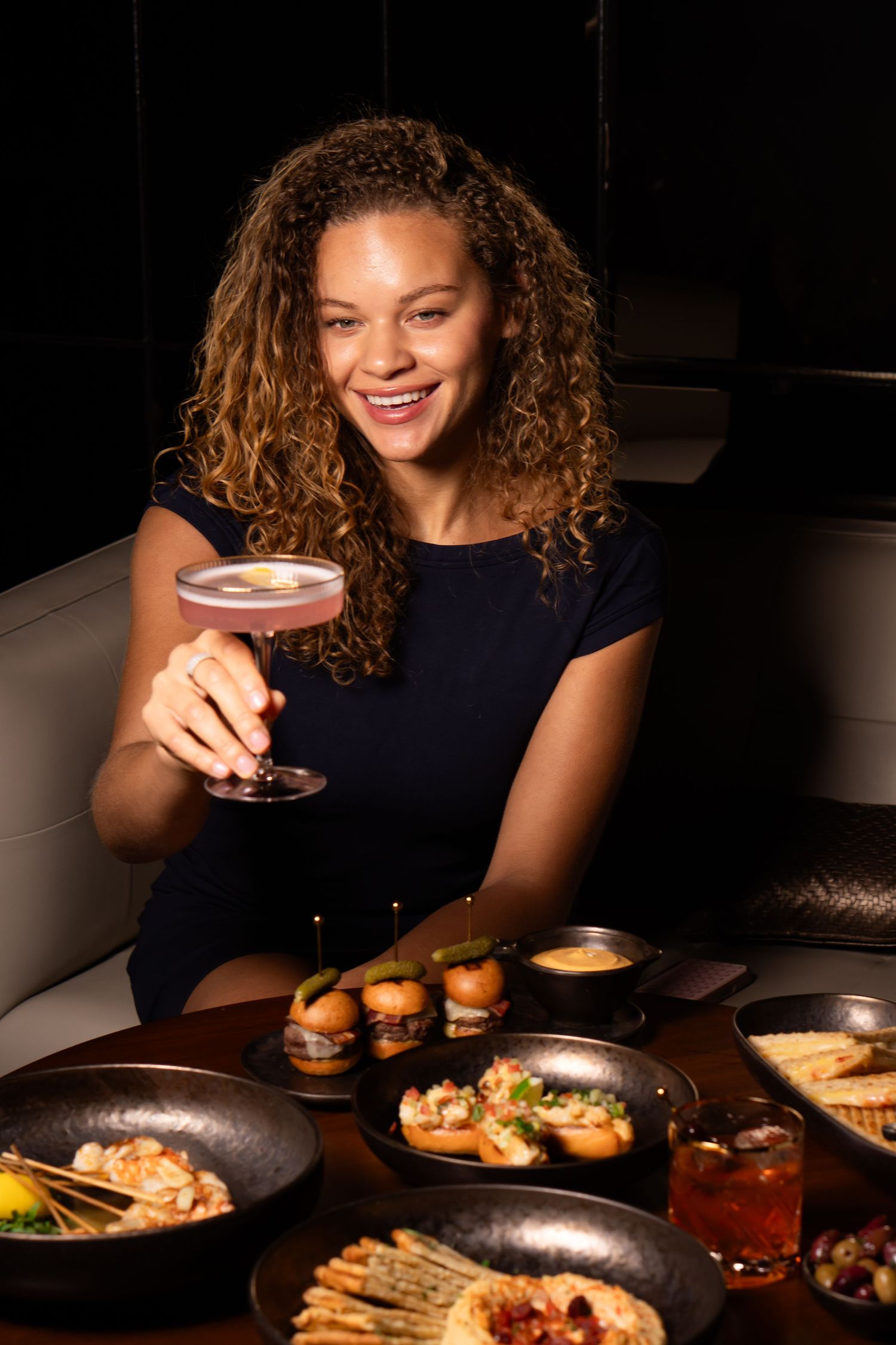 Smiling woman holding a pink cocktail at a dark restaurant table filled with elegant appetizers and drinks.