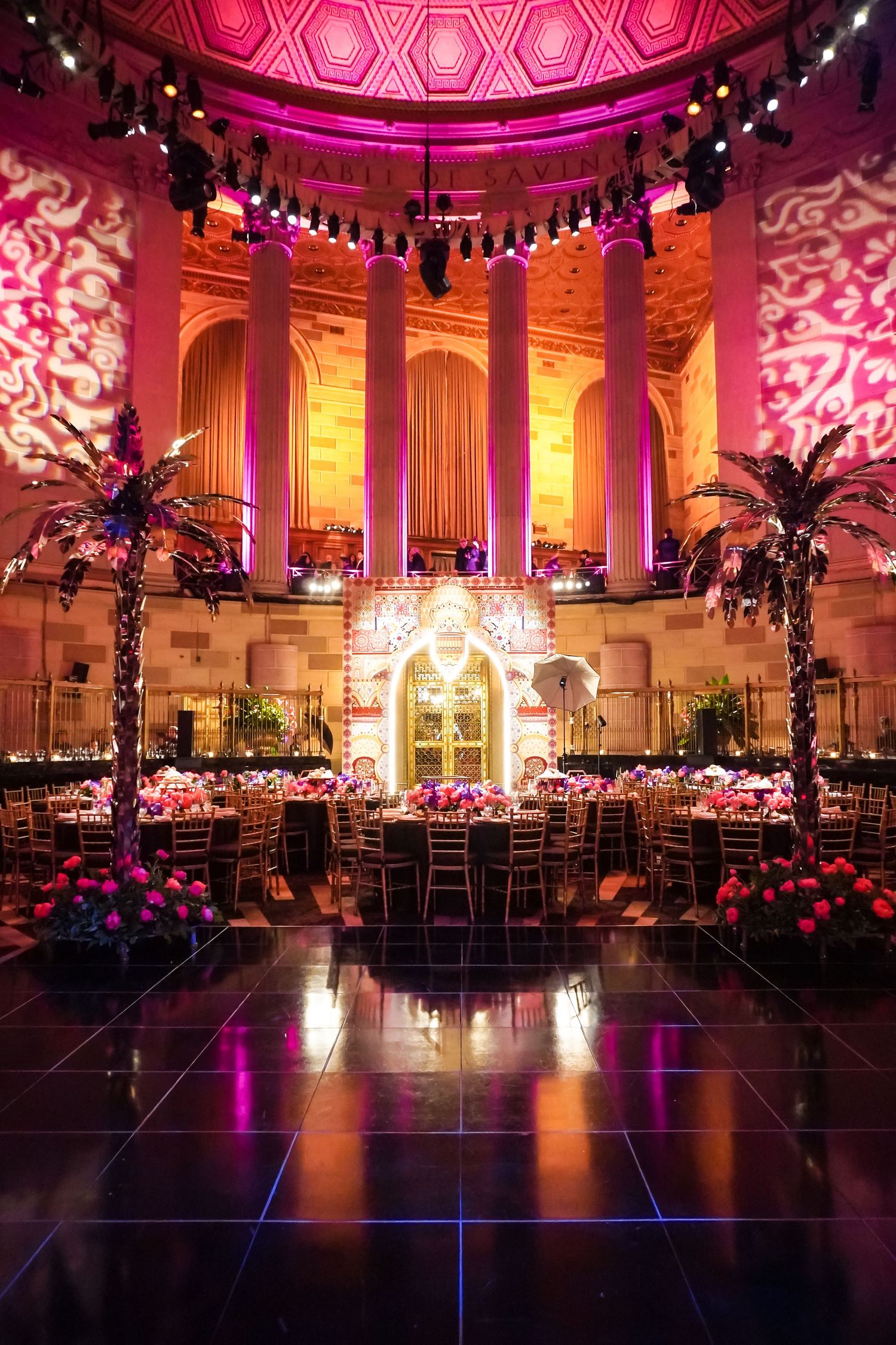 Elegant ballroom reception venue with pink uplighting, ornate columns and floral centerpieces creating a grand atmosphere.