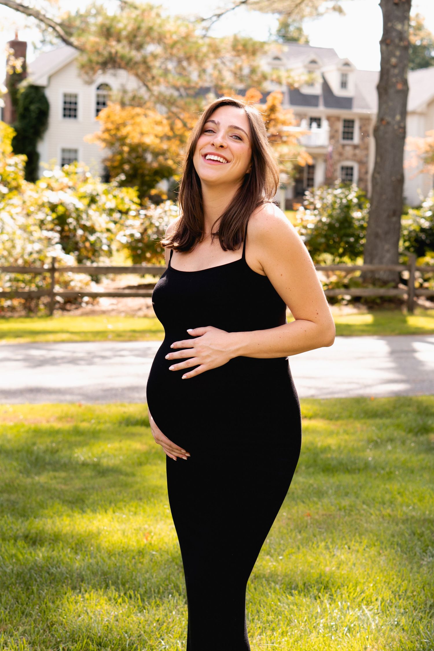 Expectant mother in black dress smiling outdoors during golden hour with trees and house in background.