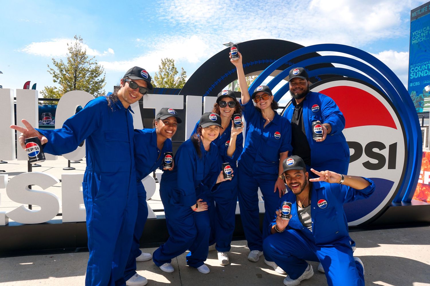 Group of people in blue jumpsuits posing in front of a Pepsi logo sign at an outdoor event.