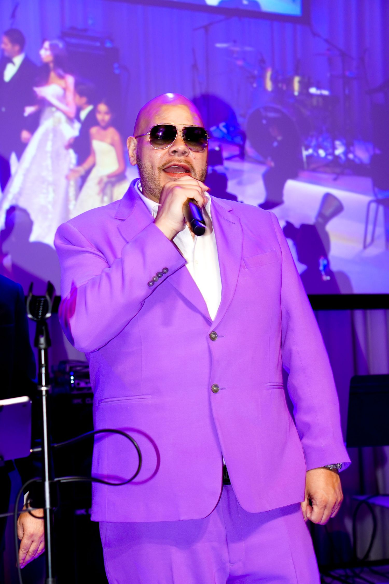 Performer in vibrant purple suit singing passionately on stage with dramatic lighting and band in background.