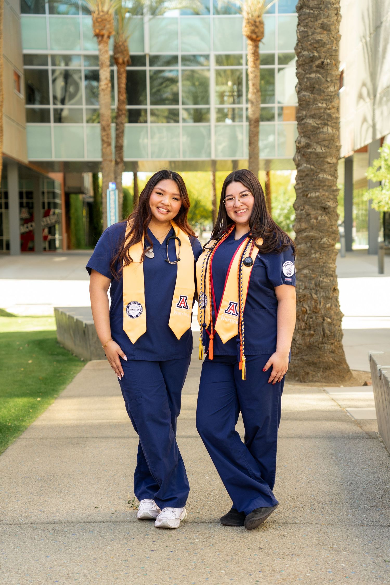 Two graduates in navy nurse uniforms and gold stoles pose together on University of Arizona walkway.