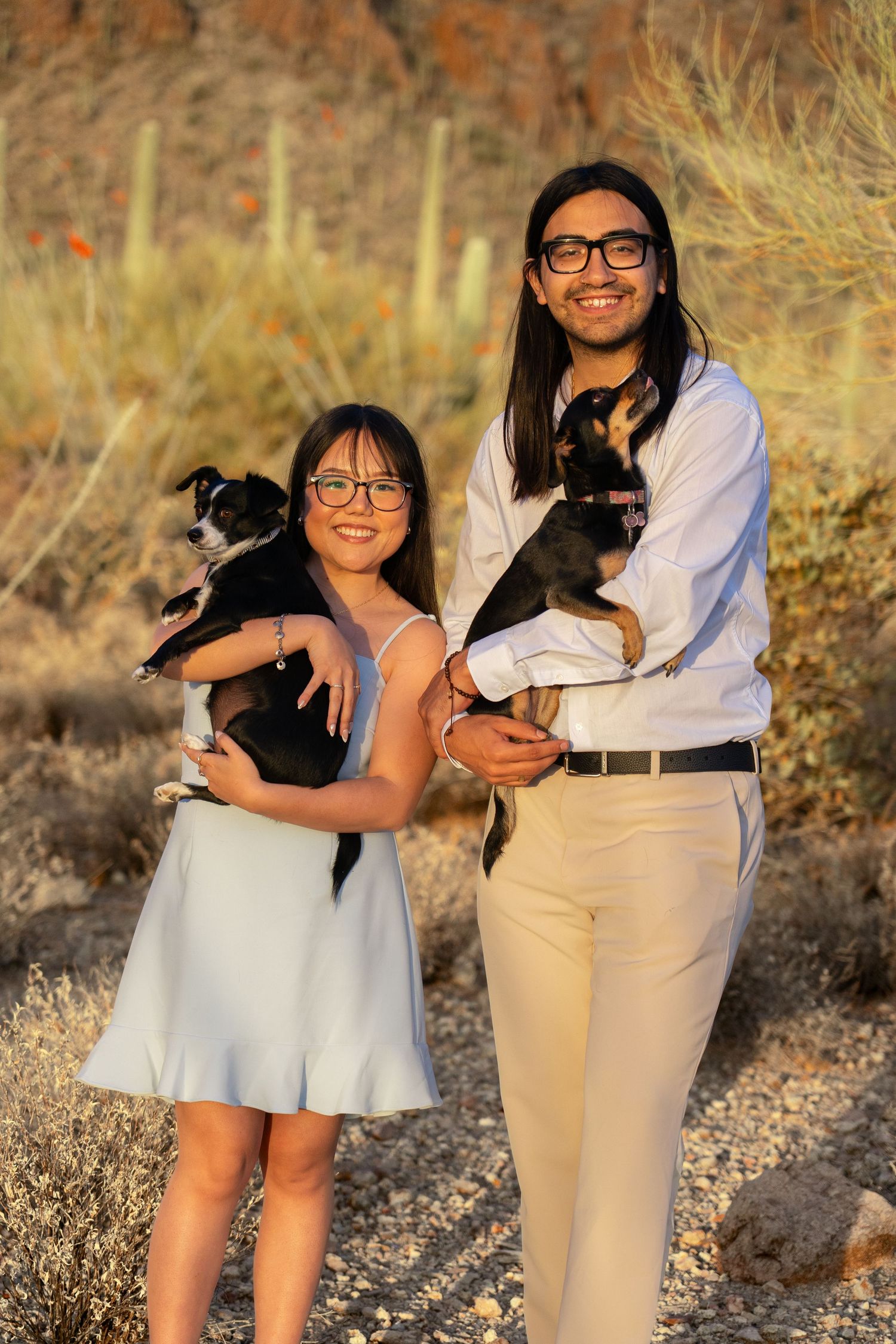 Couple in coordinated white and beige outfits pose with their small dogs against desert mountain backdrop.