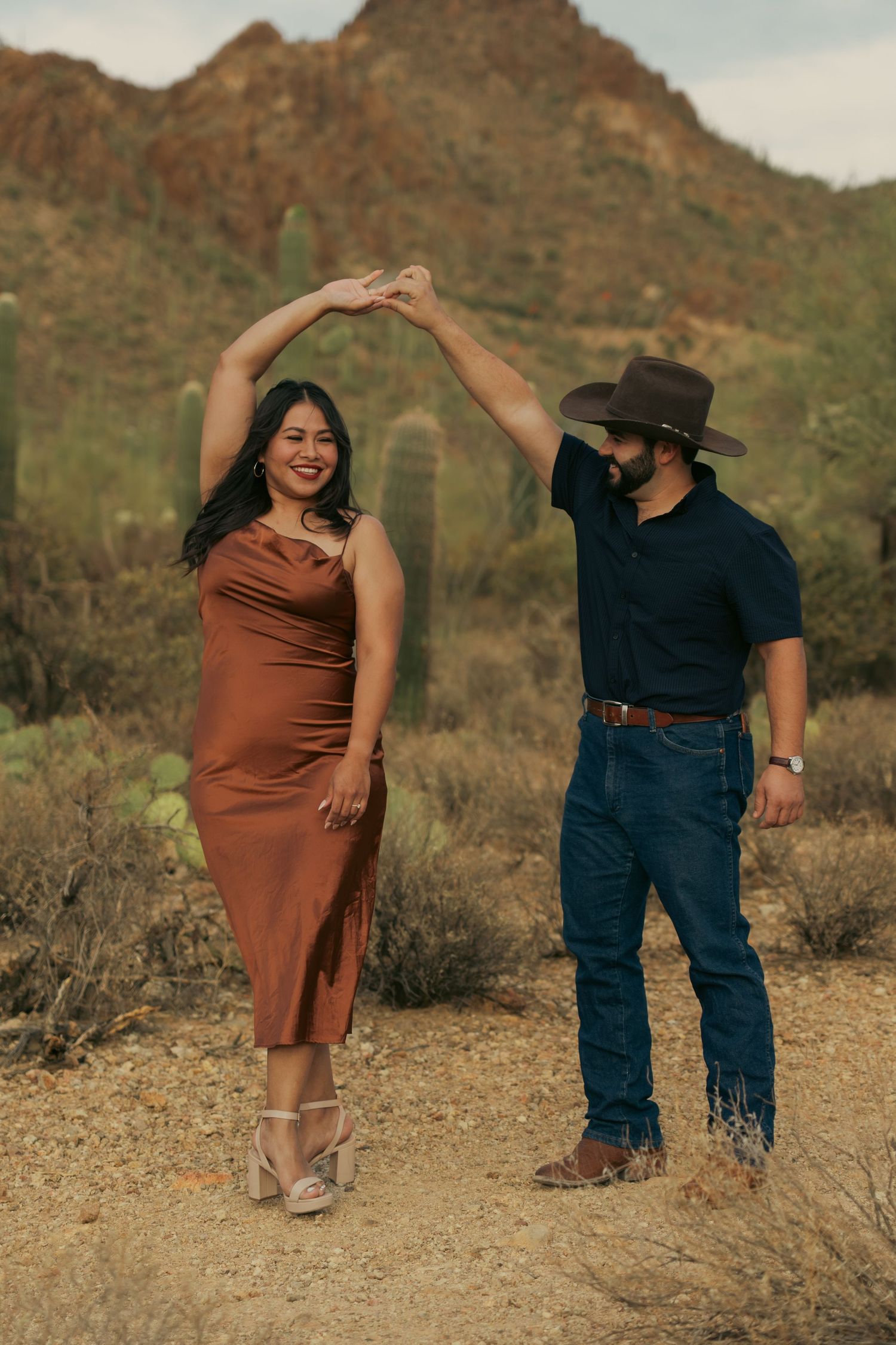 A couple in western attire dances together against a desert mountain backdrop with saguaro cacti.