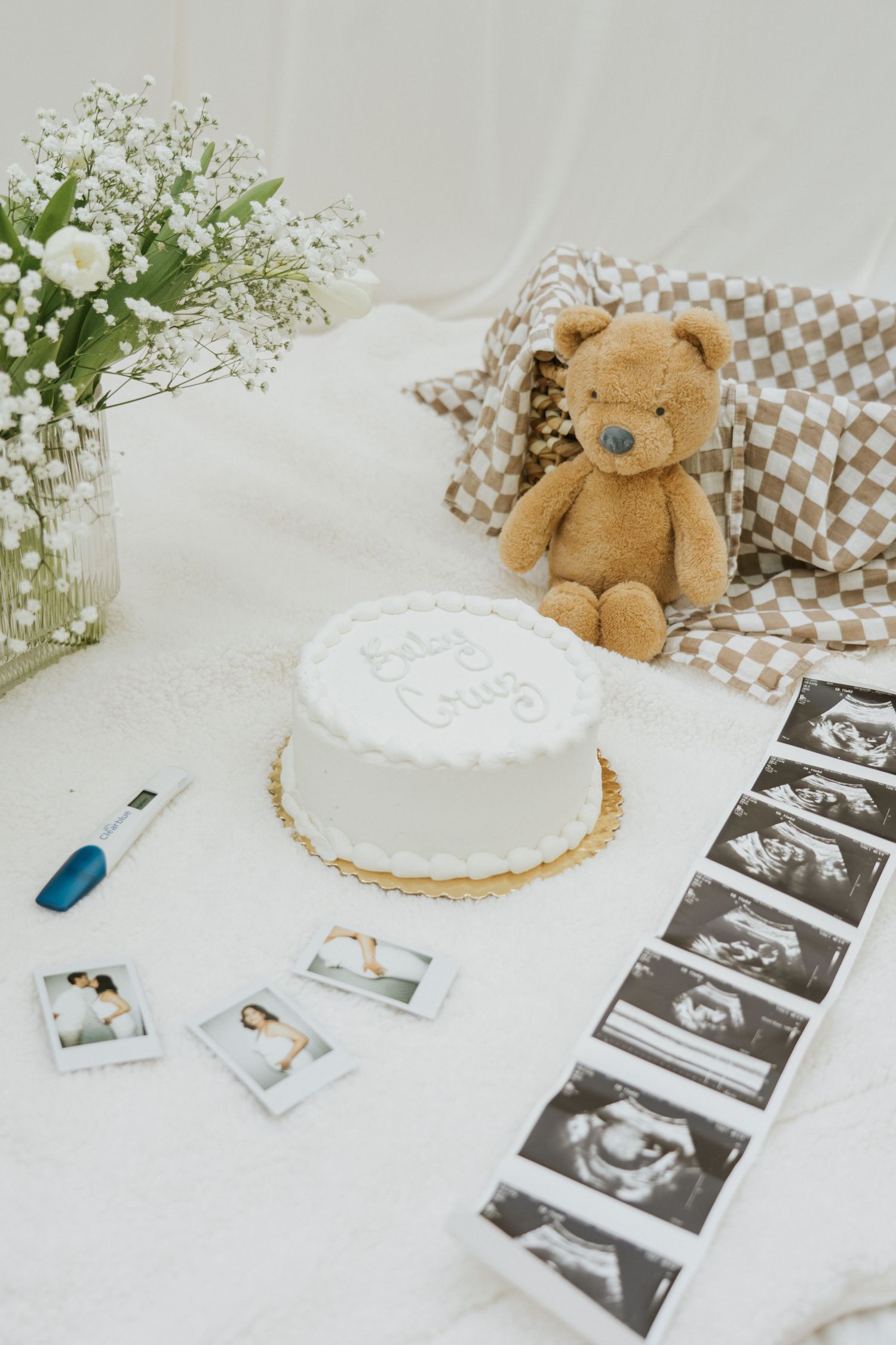 Pregnancy announcement setup with ultrasound photos, baby shoes, and delicate white flowers on a light background.