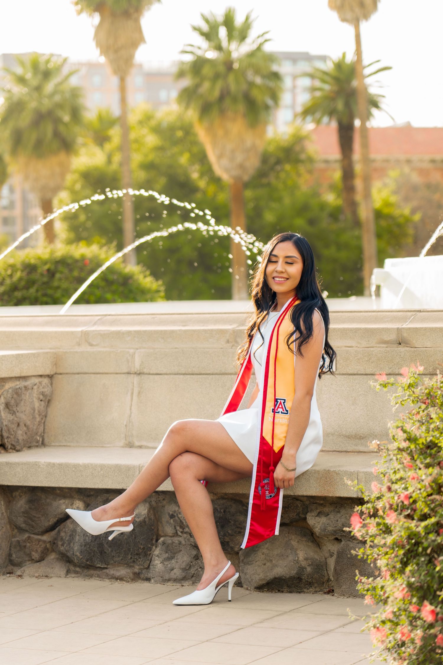 University of Arizona graduate smiles in white dress with red and gold stole, seated by fountain with palm trees.